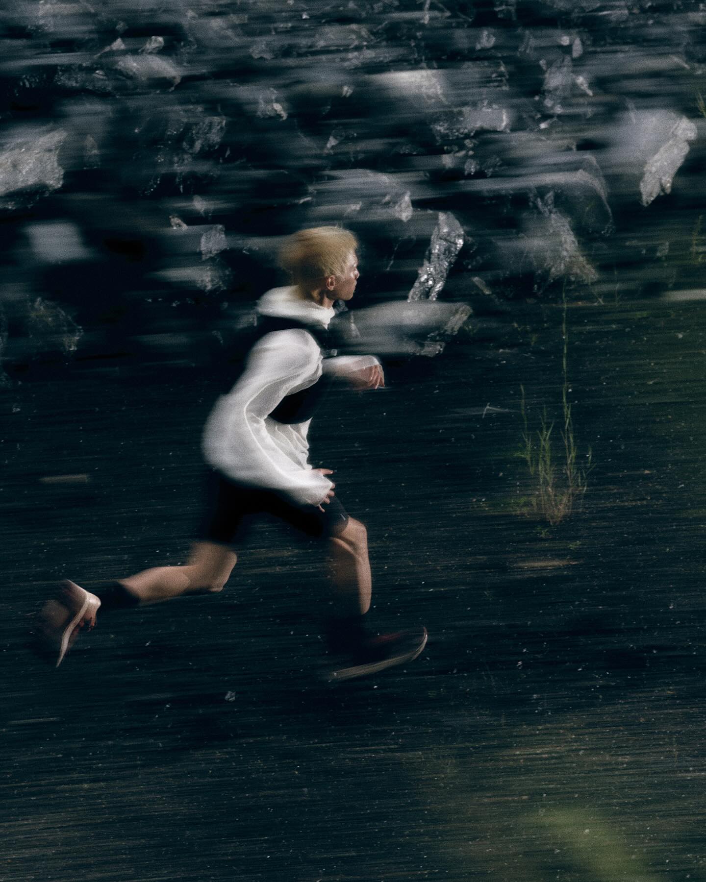 A blurred figure in a white jacket and black shorts runs across a dark, textured ground against a backdrop of jagged, icy rocks.