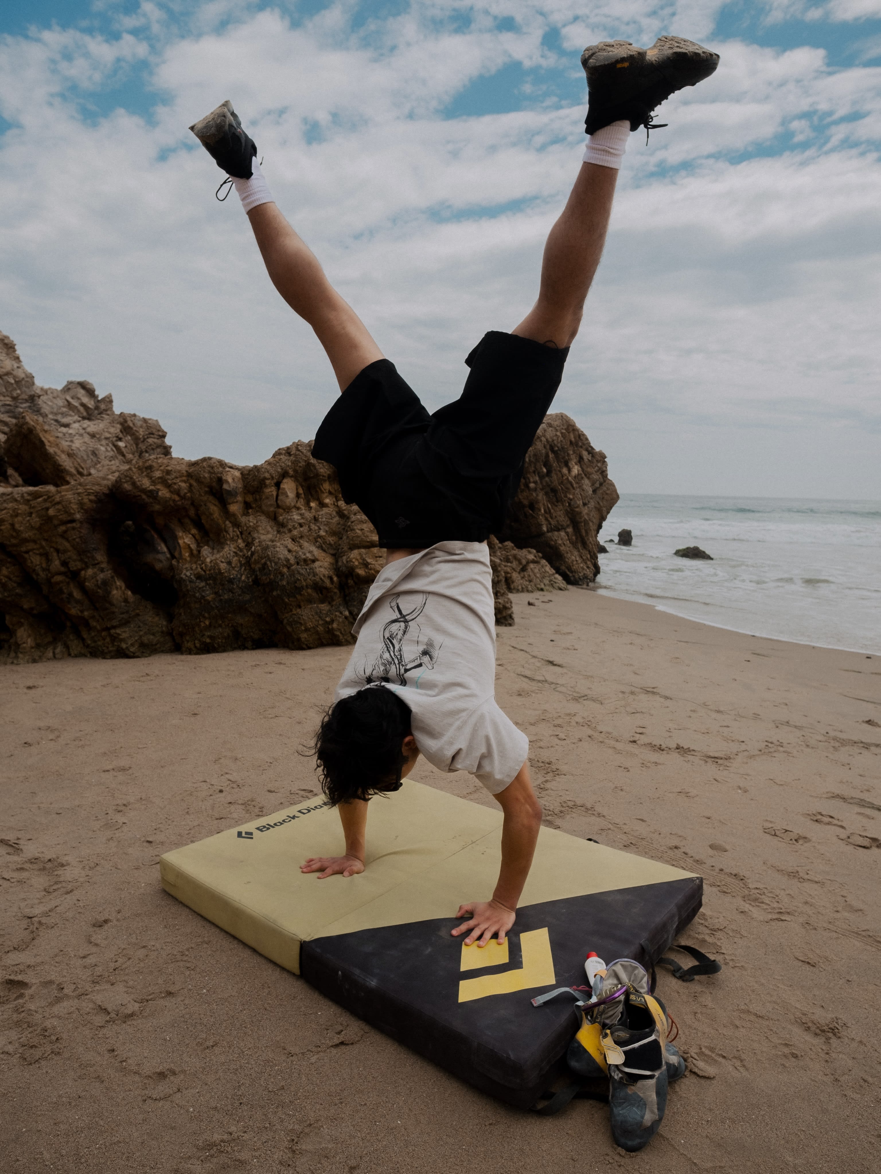 A person performs a handstand on a crash pad on a sandy beach, with rocky cliffs and a cloudy sky in the background.