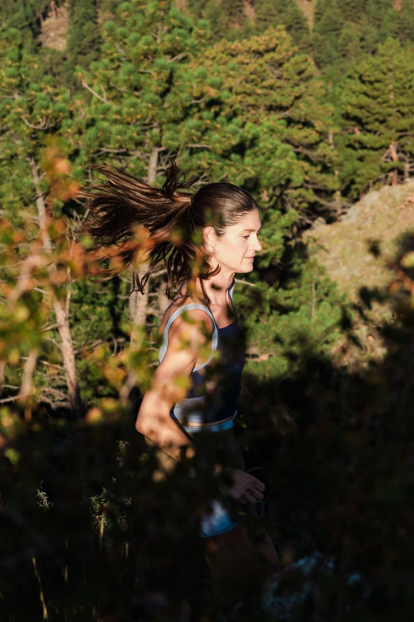 A woman with flowing hair runs through a brushy foreground against a backdrop of dense green pine trees.