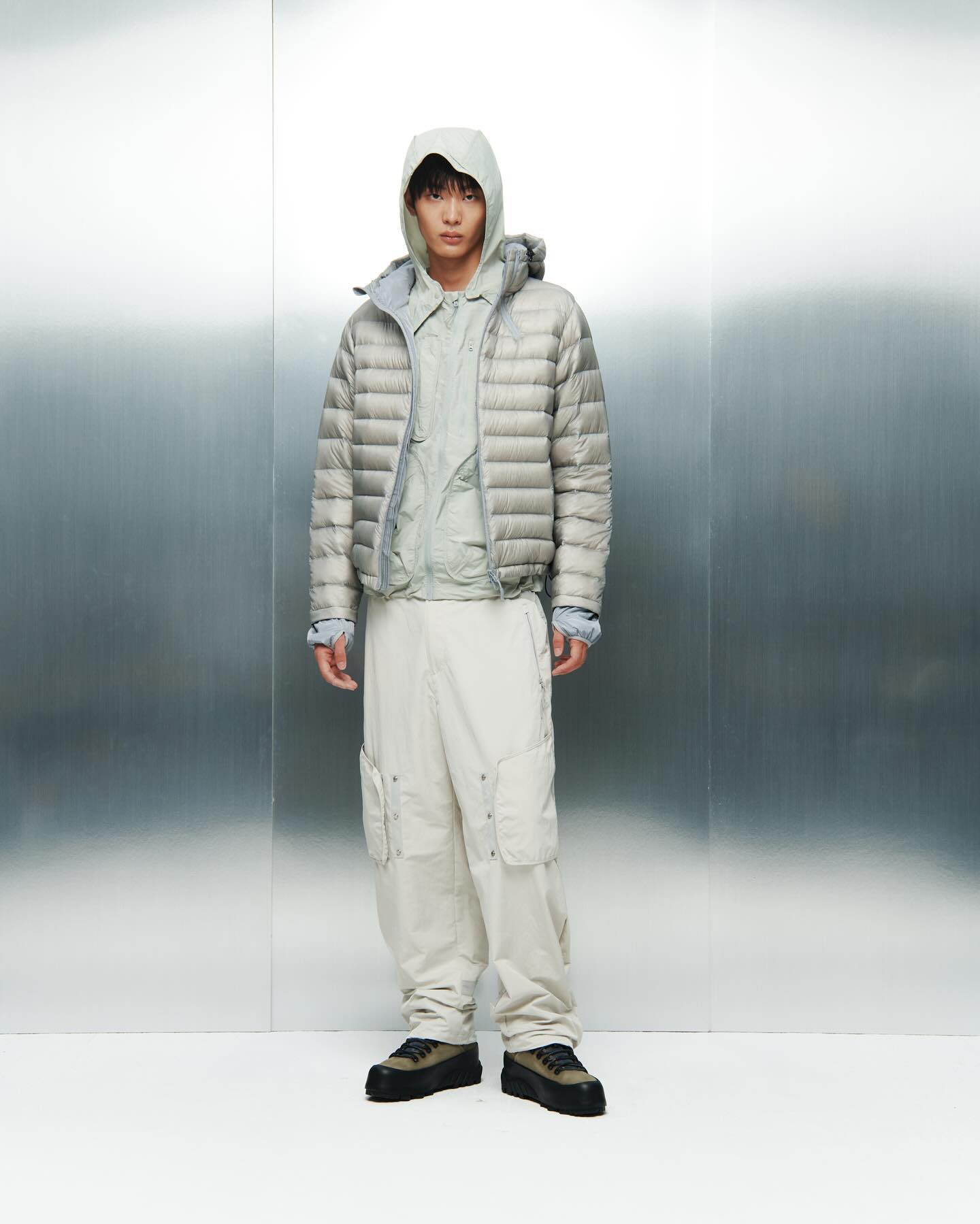 A man wearing a light-colored puffer jacket, hoodie, and cargo pants stands against a metallic silver wall.