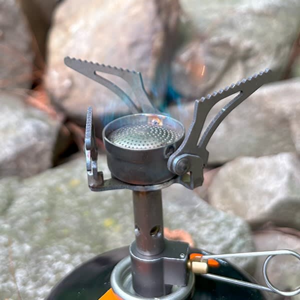 A small camping stove with a blue flame sits atop a fuel canister against a blurred background of grey rocks.