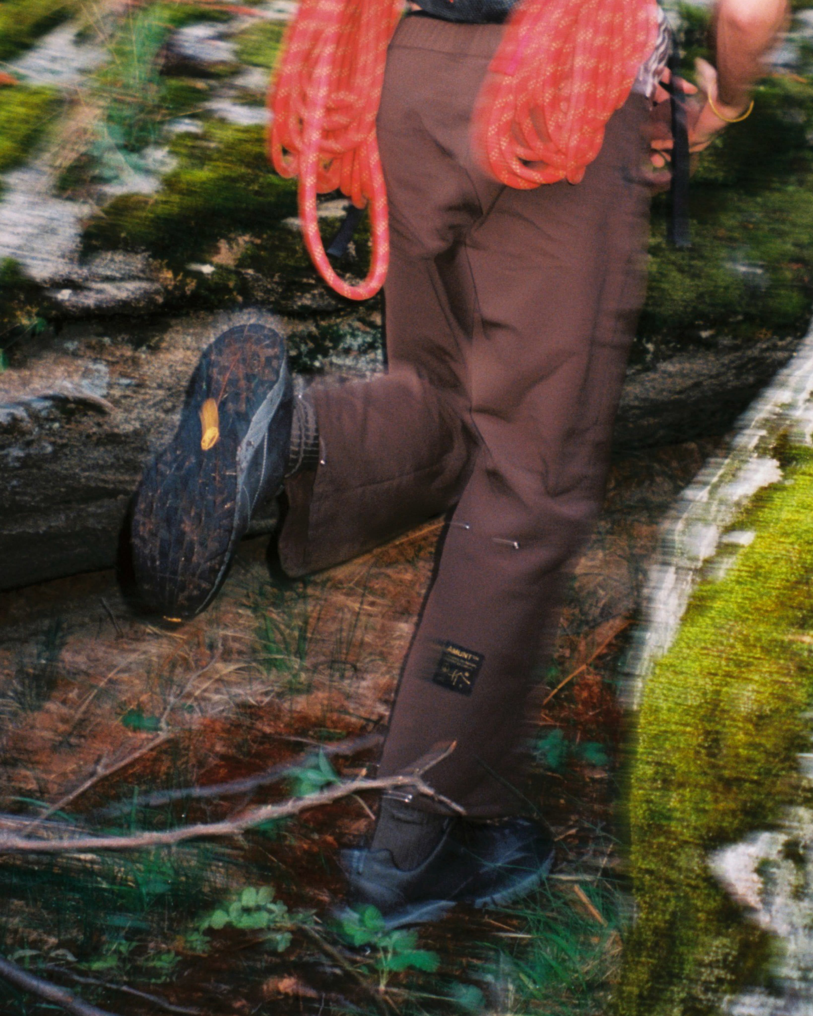A person wearing brown pants and hiking boots stands on a forest floor with a coil of orange rope around their waist.