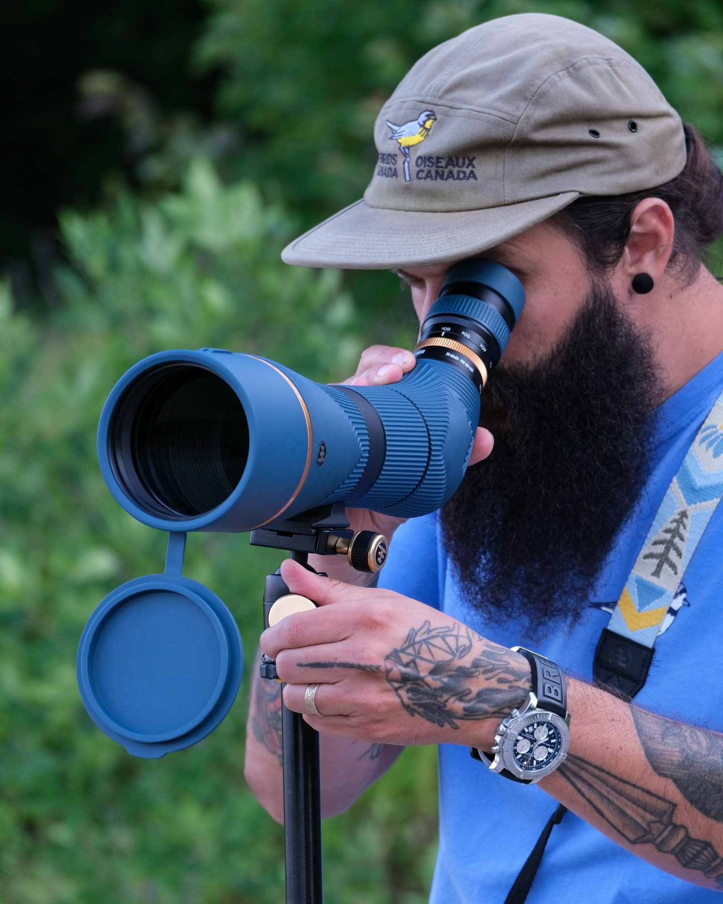 A man with a beard and tattoos, wearing a cap, looks through a spotting scope in the foreground, with blurred green foliage in the background.