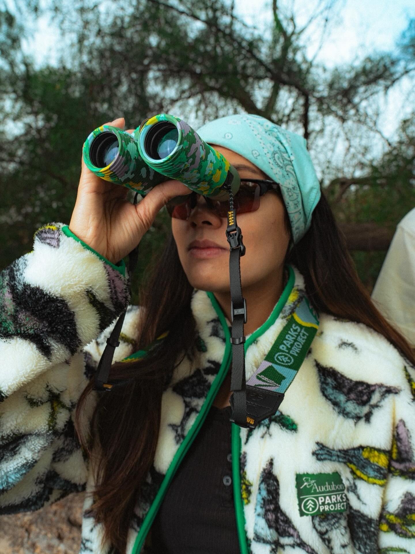 A woman in a bird-patterned fleece jacket and bandana looks through camouflage binoculars in a wooded area.