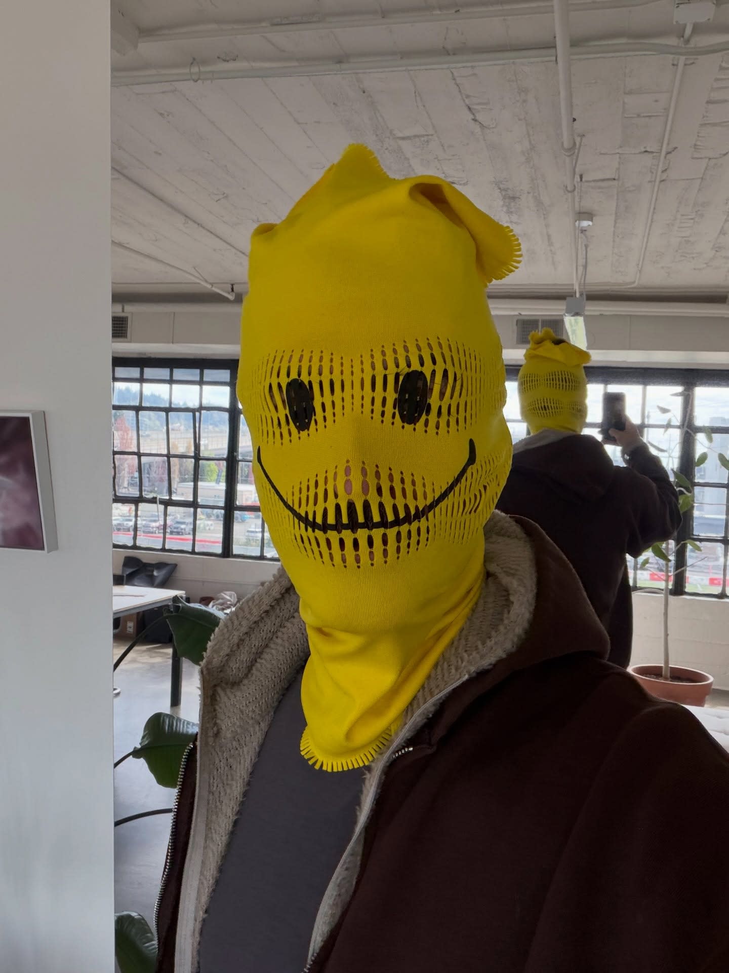 A person wearing a yellow knitted smiley-face mask and a brown jacket stands in a bright room with large windows and a white ceiling.
