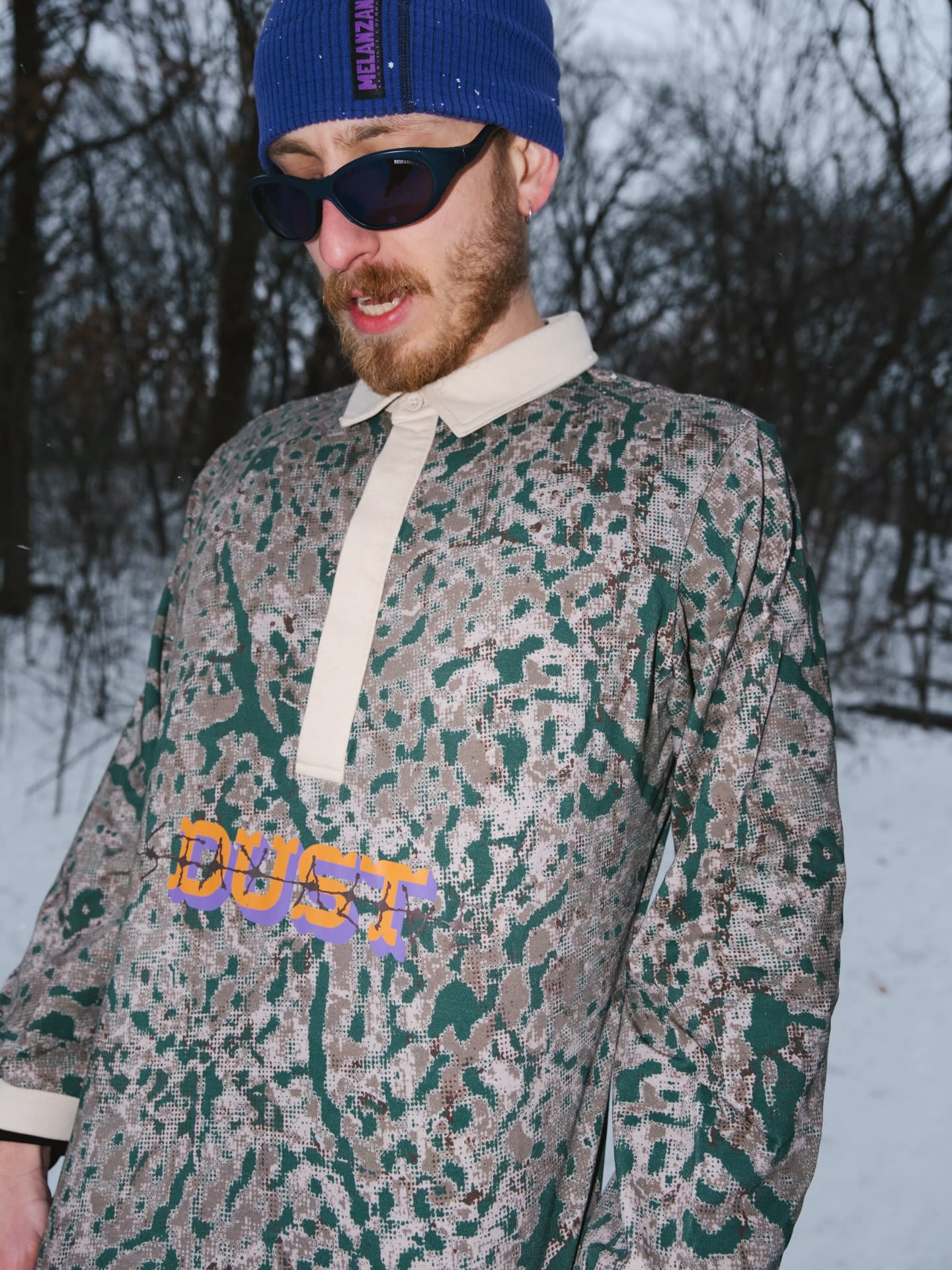 A man wearing a blue beanie, sunglasses, and a camouflage-patterned shirt stands against a snowy backdrop of bare winter trees.