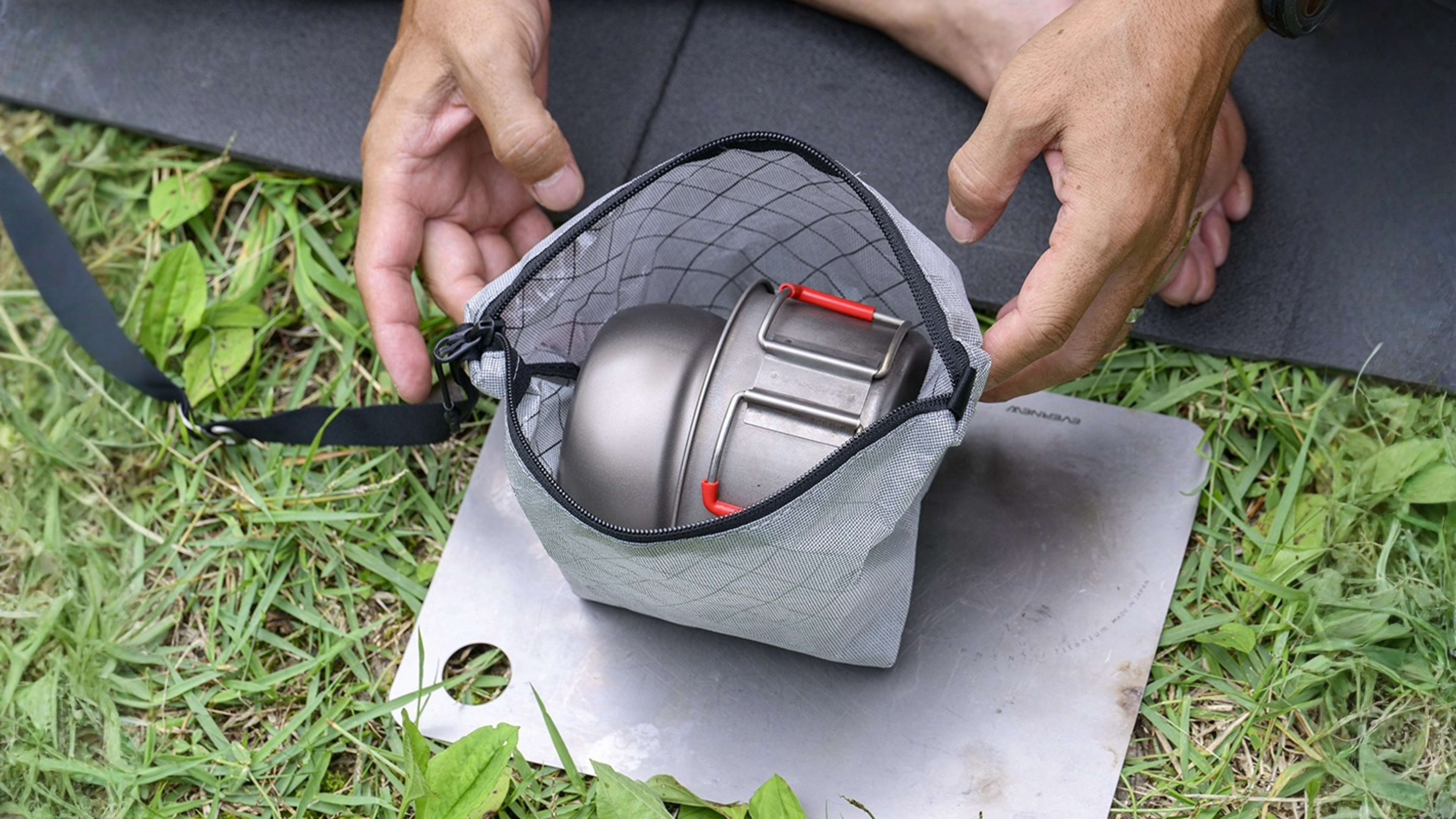 A person's hands hold open a grey mesh pouch containing a metal camping pot, resting on a silver tray over green grass and a black mat.