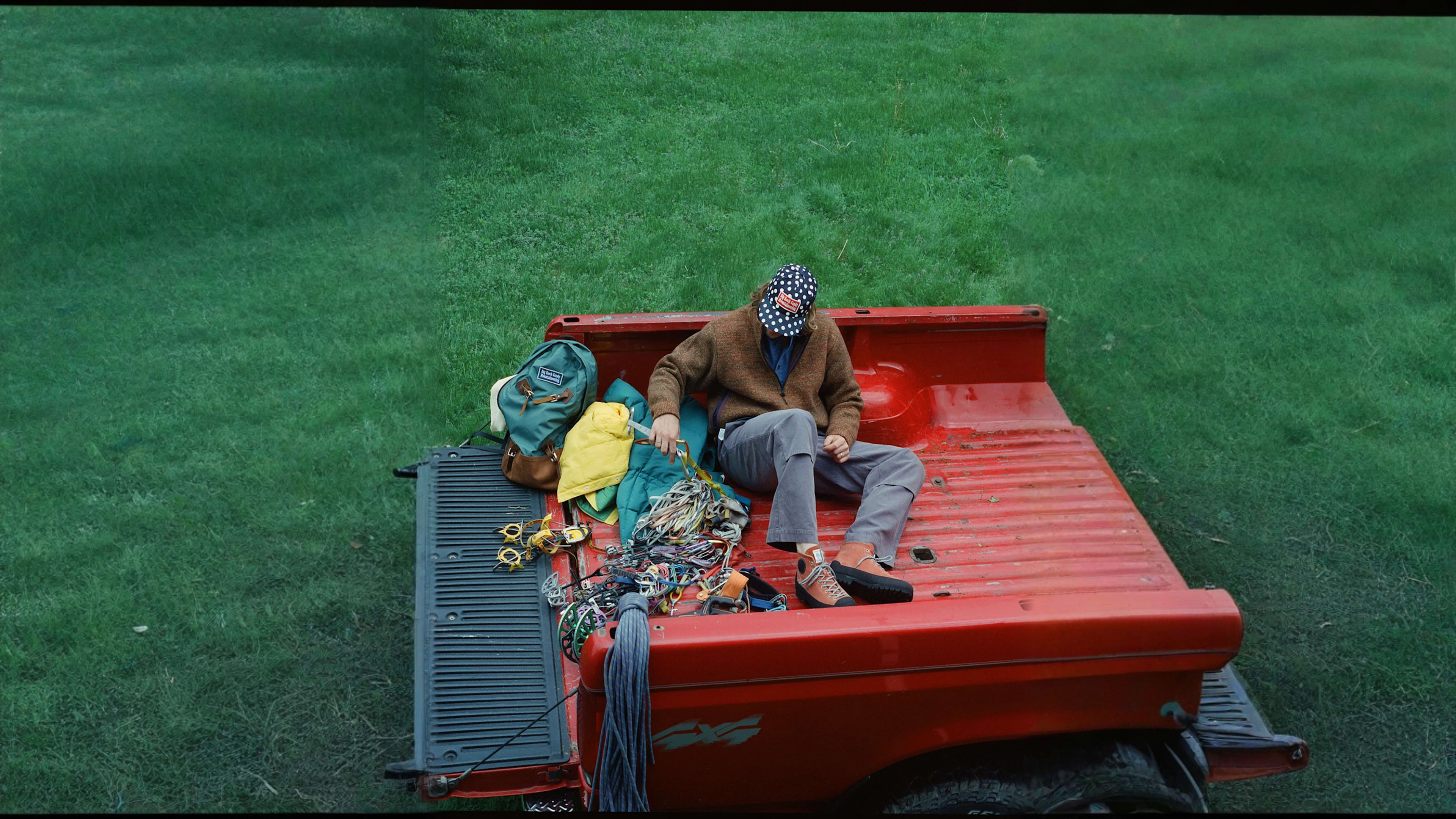 A person wearing an American flag hat sits in the bed of a red pickup truck filled with climbing gear, with a lush green lawn in the background.