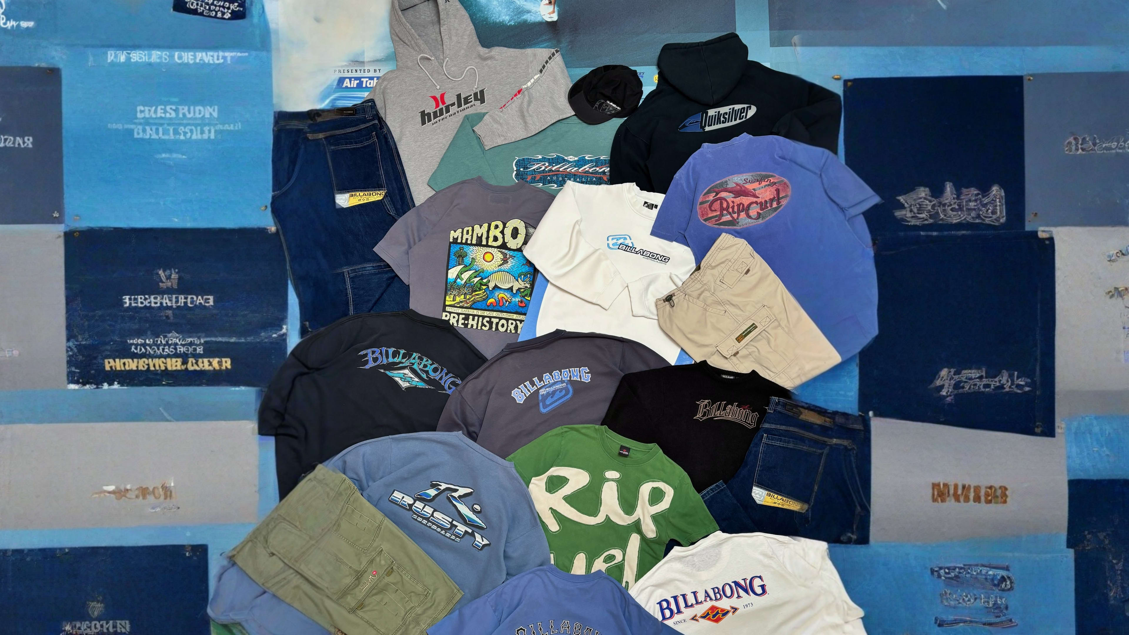 A variety of surf-brand hoodies, t-shirts, and pants are spread across a blue surface.