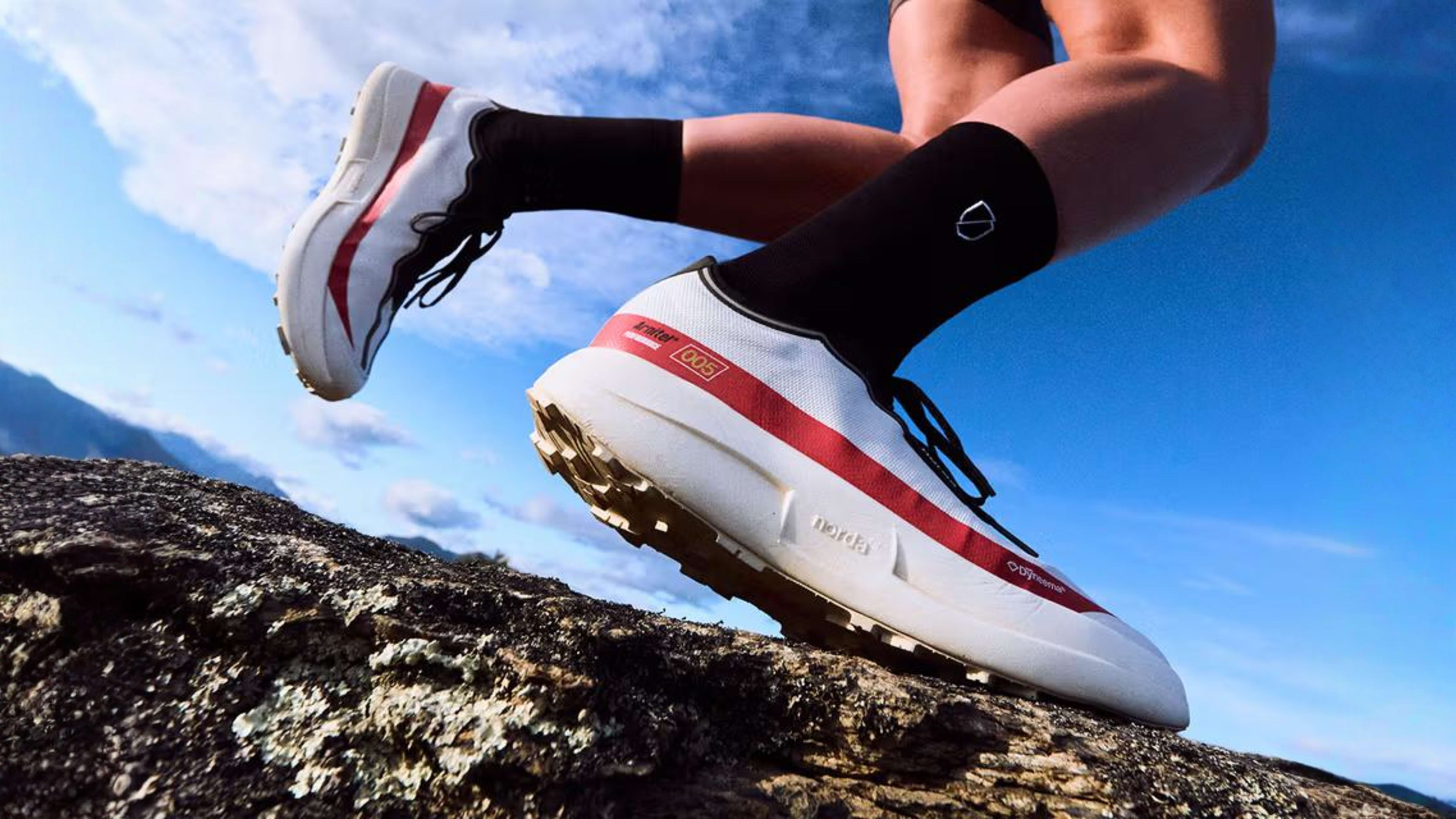 Feet in white and red trail running shoes stepping on rocky terrain under a blue sky.