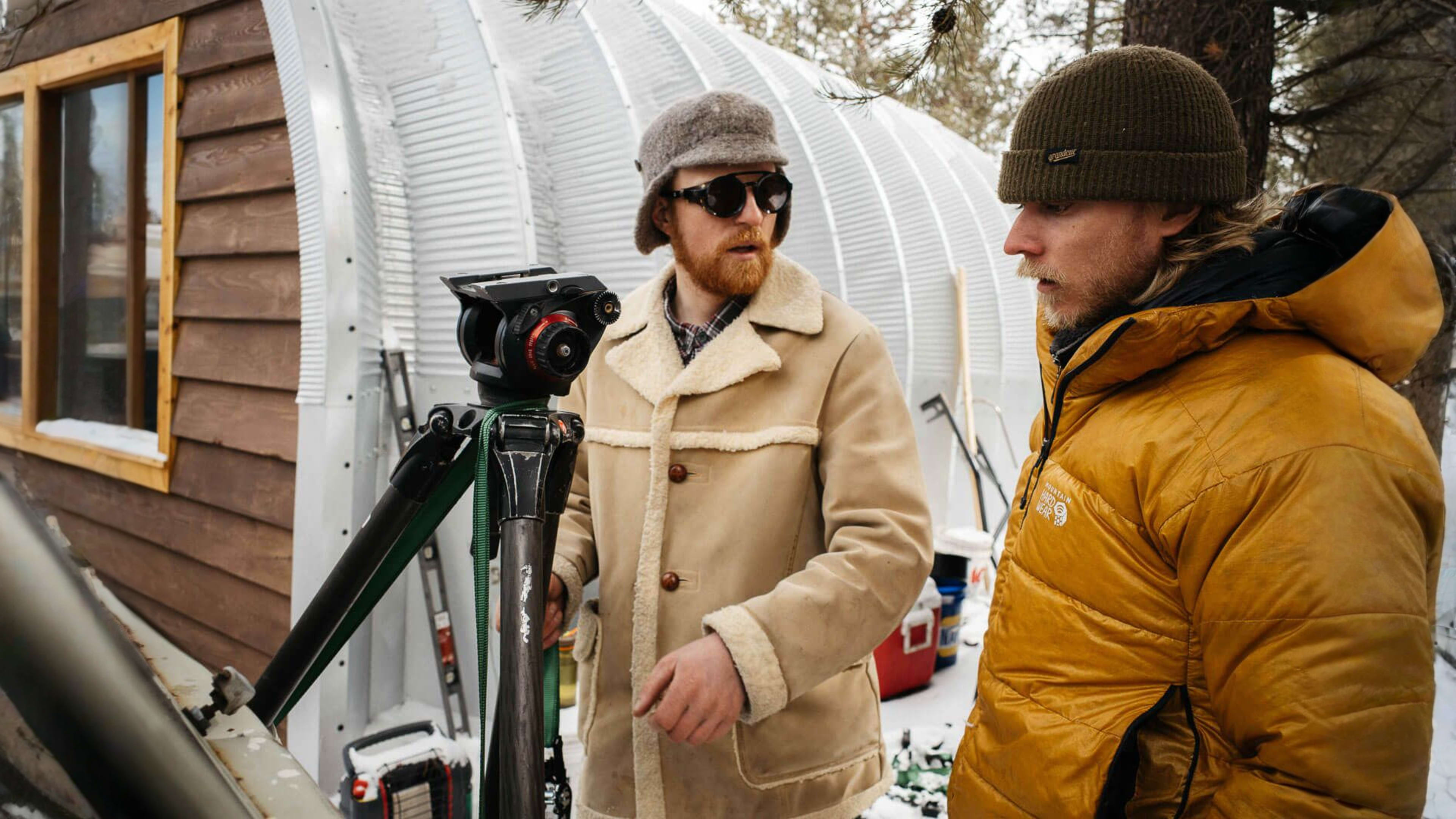 Two men in winter clothing, one with a camera on a tripod, talk in a snowy outdoor setting with buildings behind them.