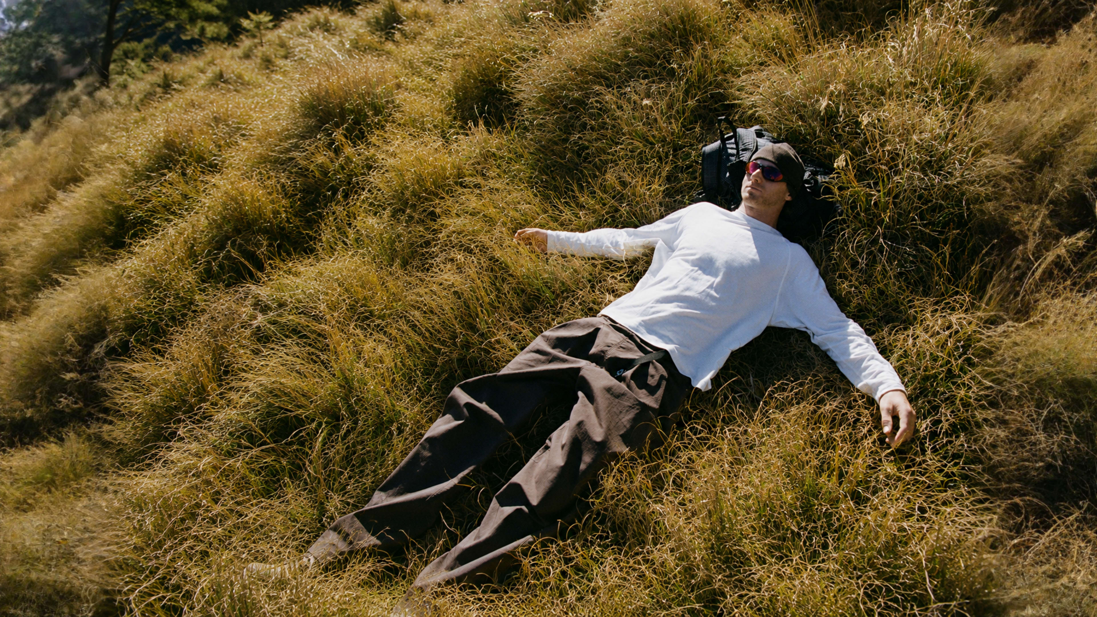 A man in a white shirt, beanie, and sunglasses rests on his back in dry, golden grass, using a backpack as a pillow.