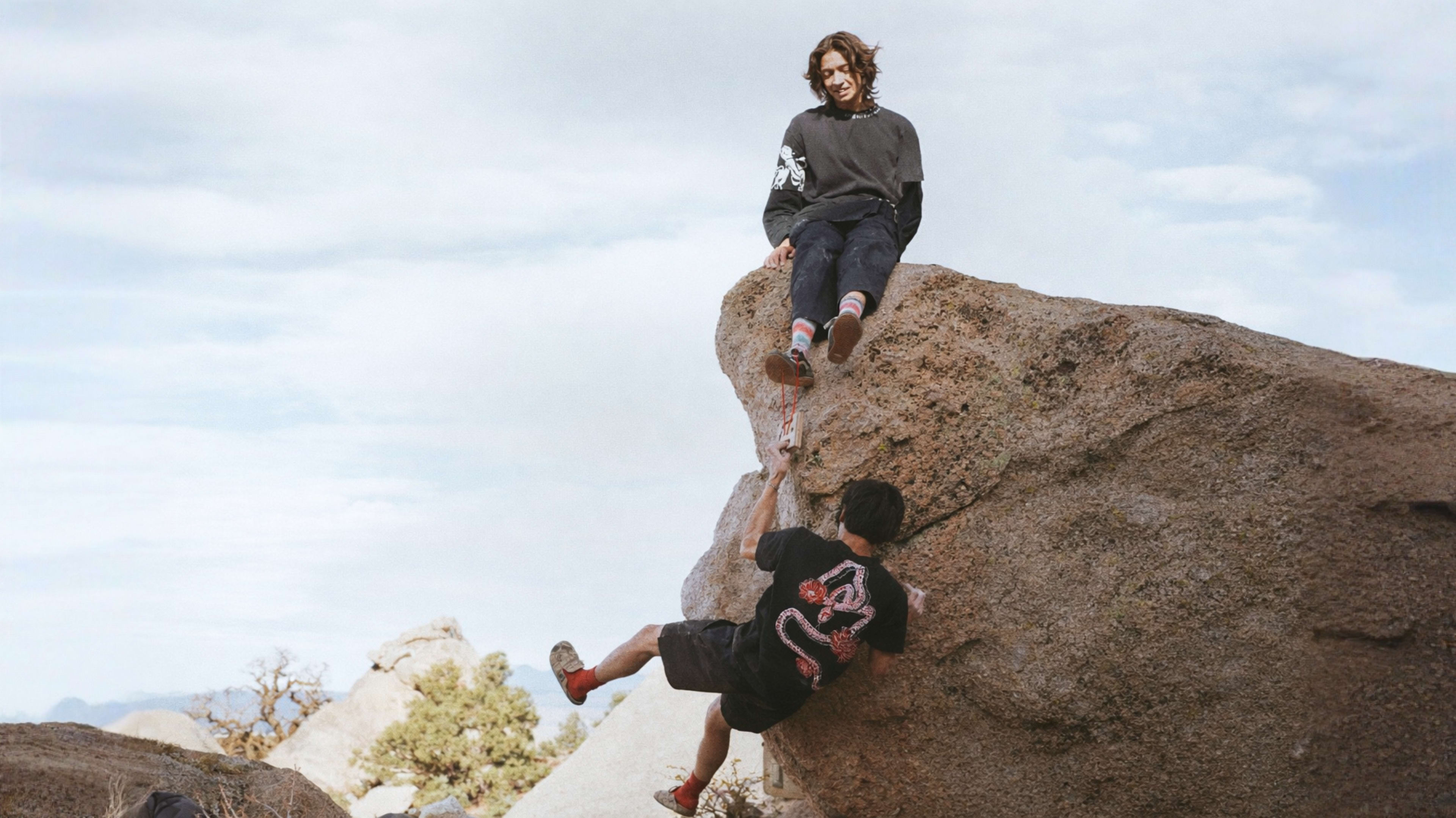 A person atop a boulder extends a hand to help another person climb up.