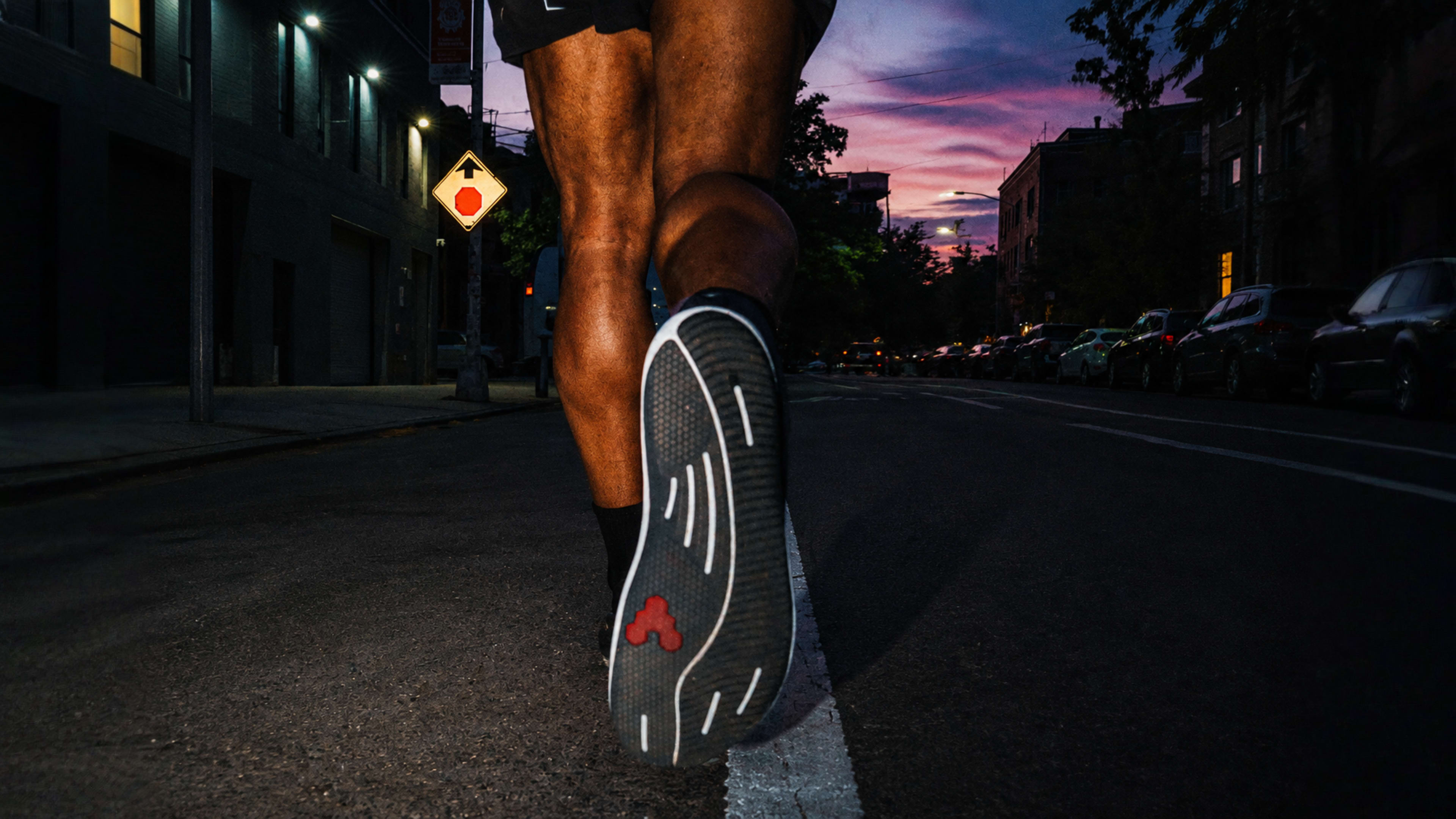 The sole of a running shoe and a person's leg mid-stride on an urban street at dusk, beneath a vibrant sky.