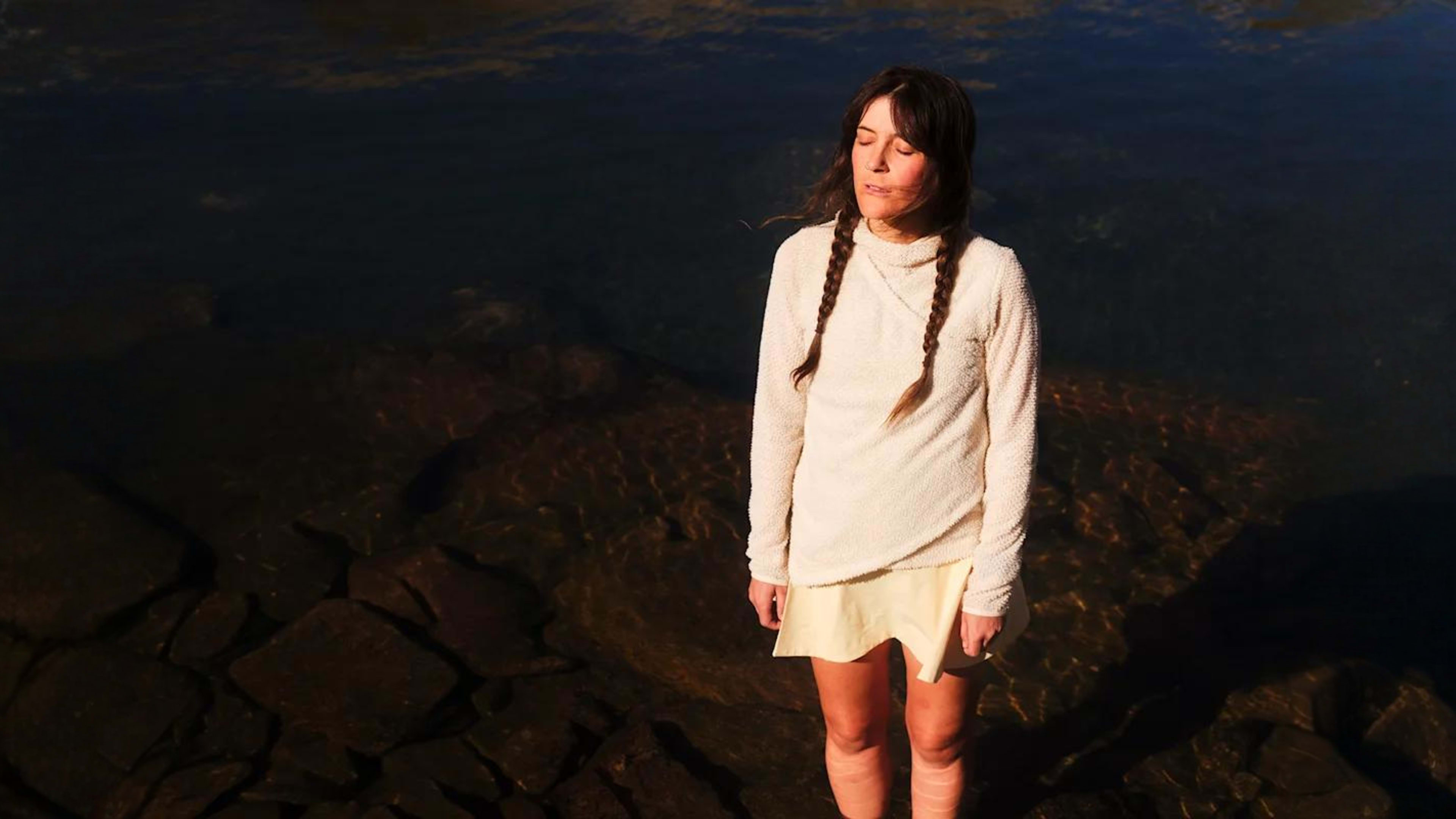Woman with two braids stands in shallow water, eyes closed.