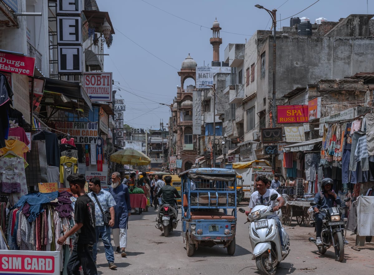 A busy shopping street in Delhi lined with shop signs, clothing stalls, an e-rickshaw and scooters.