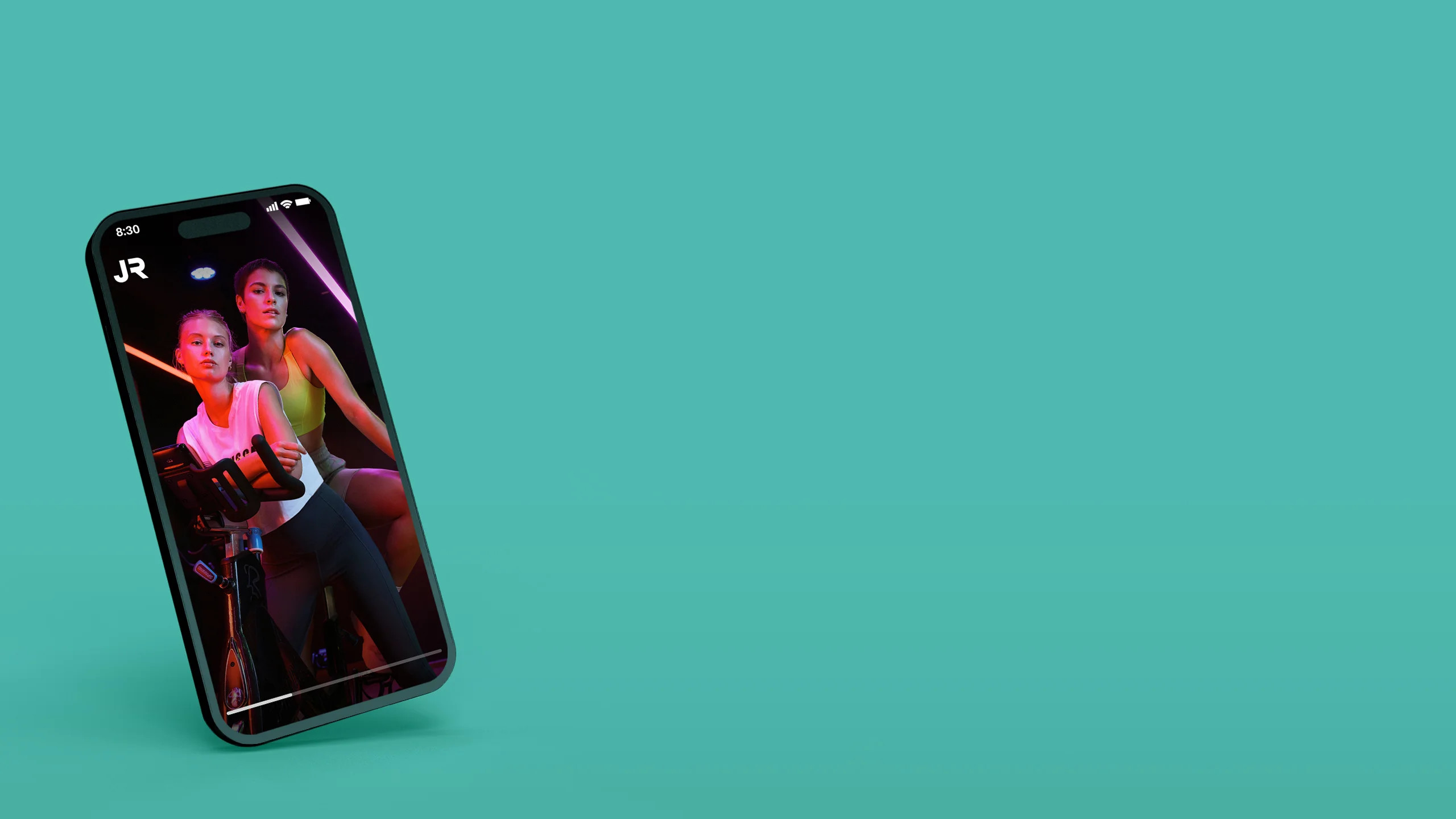 Smartphone links vor grünem Hintergrund, auf dem Bildschirm sind zwei Sportlerinnen auf einem Indoor Bike zusehen, das John Reed Logo ist oben links auf dem Bildschirm des Telefons sichtbar. 