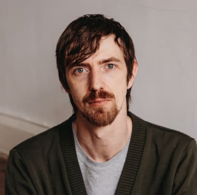 Liam Merlyn wearing a green cardigan and grey t-shirt, looking at the camera with a neutral expression.