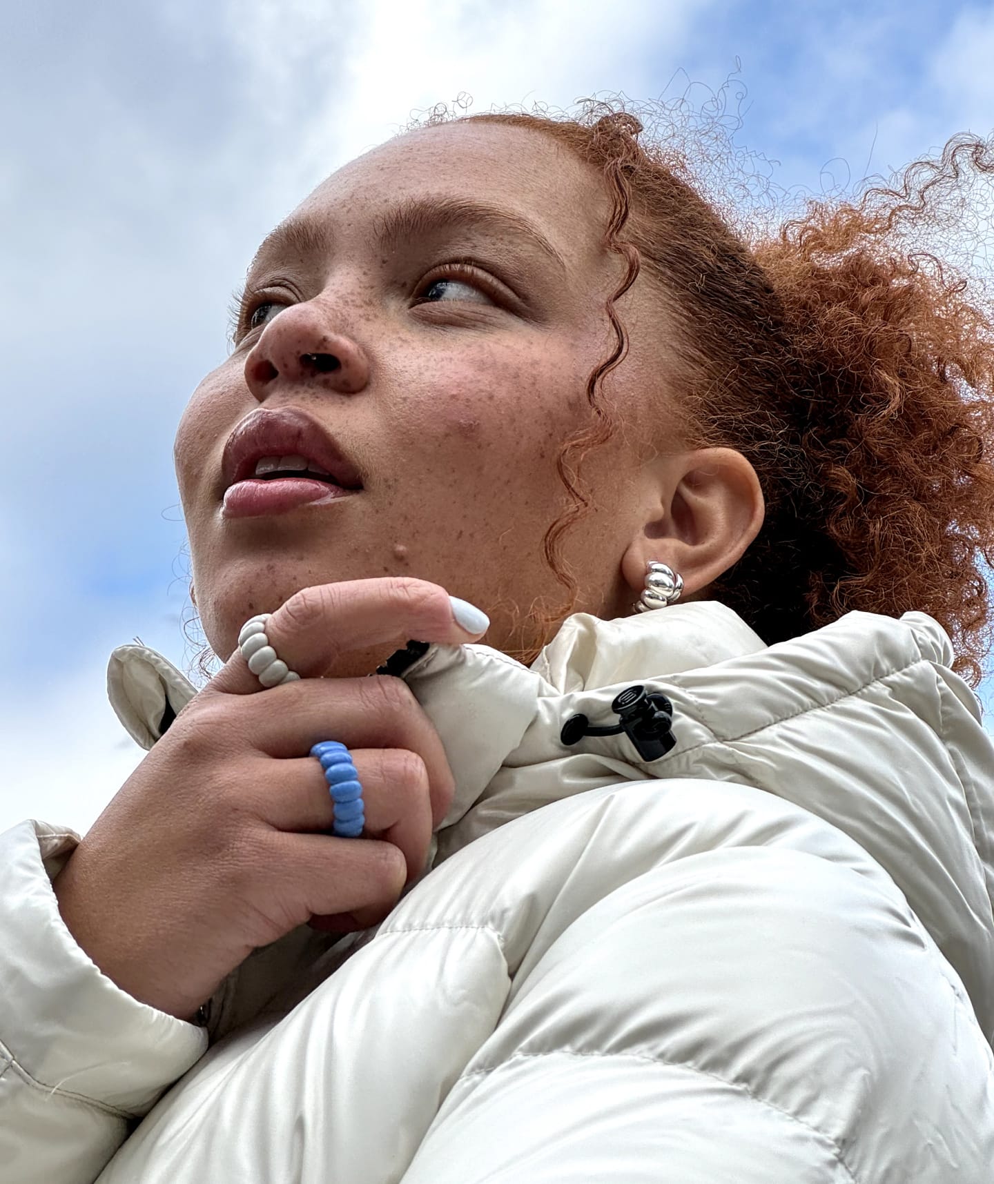 Model is wearing the Cloud and Sky Enamel coloured puffy charlotte rings while holding her whie puffer coat outside looking at the sky. 