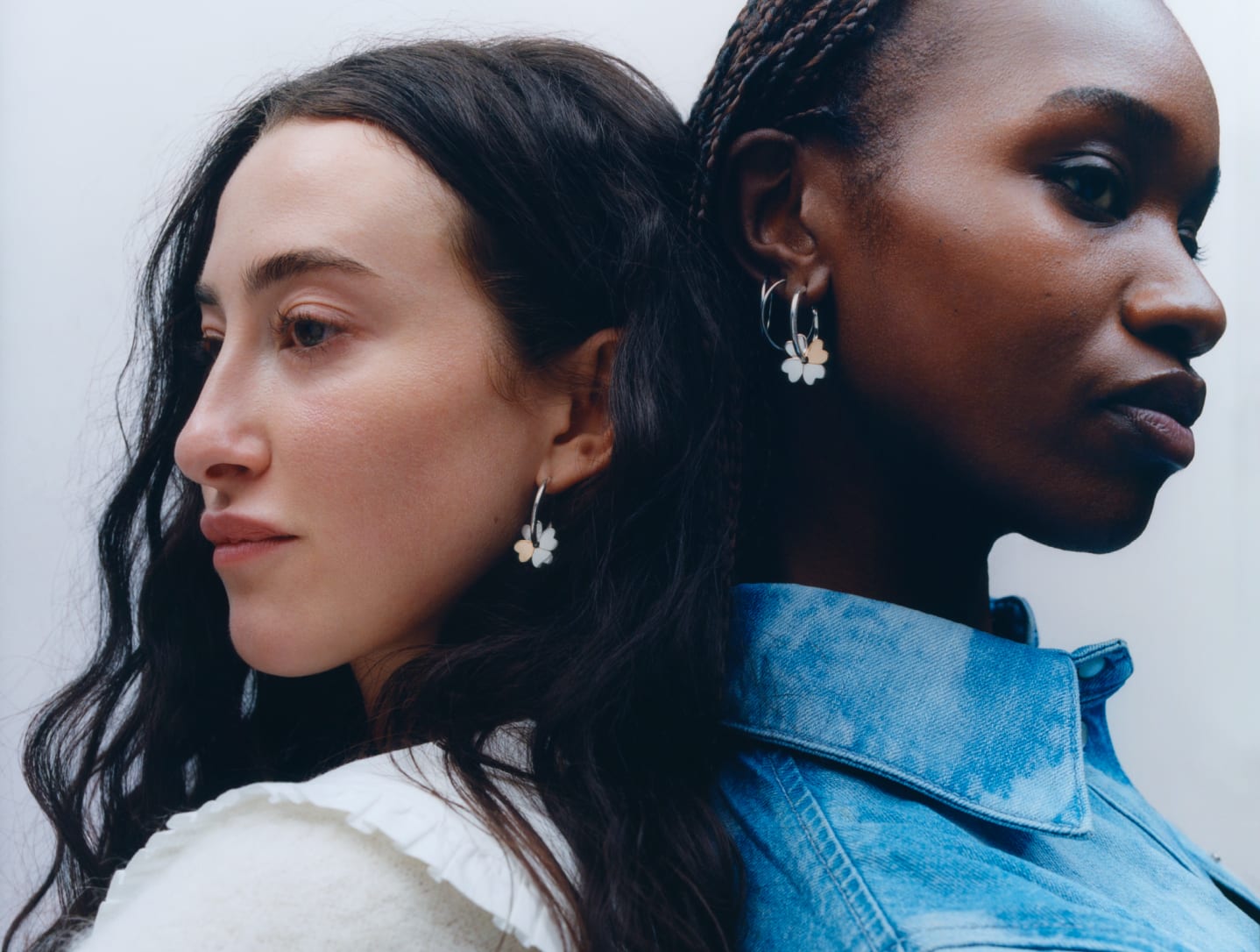 Tonia and Karla are standing back to back. They are wearing matching Mejuri x Ganni Clover hoop earrings