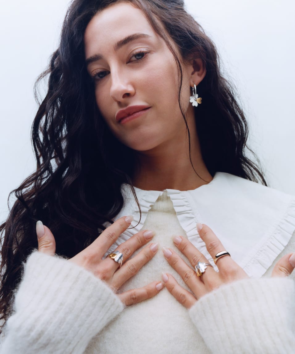 Mode is wearing a frilly oversized peter pan collar white shirt, with a fuzzy white sweater. Her hands are on her collar bones and she has Mejuri x Ganni mixed metal clover rings on her fingers.