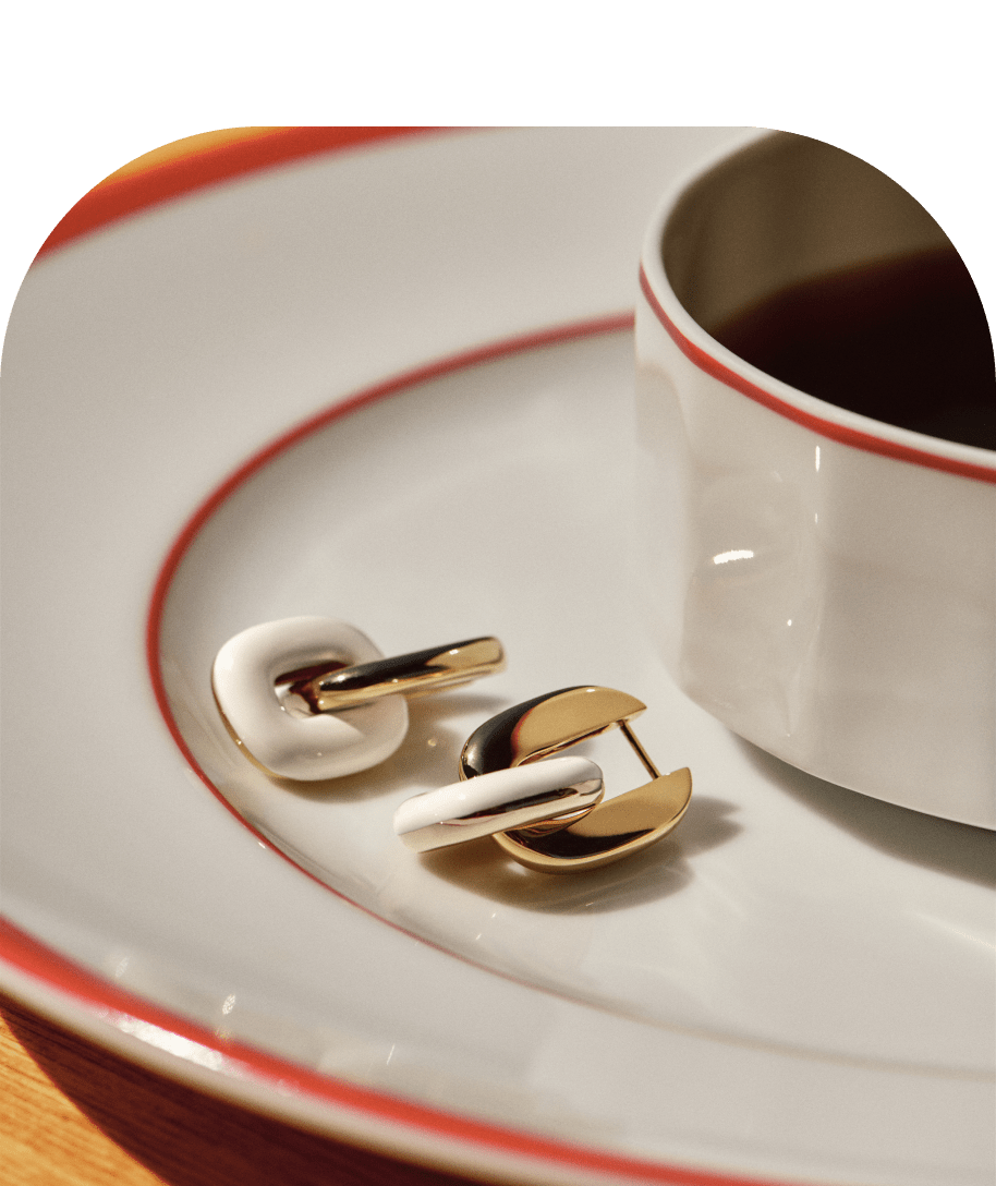 The Stevie Hoops in Cream Enamel are resting on a saucer with a cup coffee. 