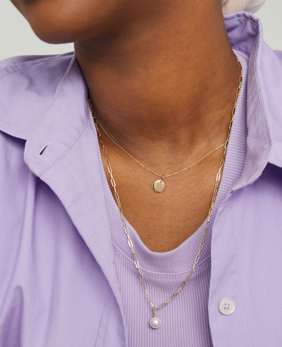 Model is wearing a lilac top featuring the Pearl Drop Charm stacked with other Mejuri necklaces and charms