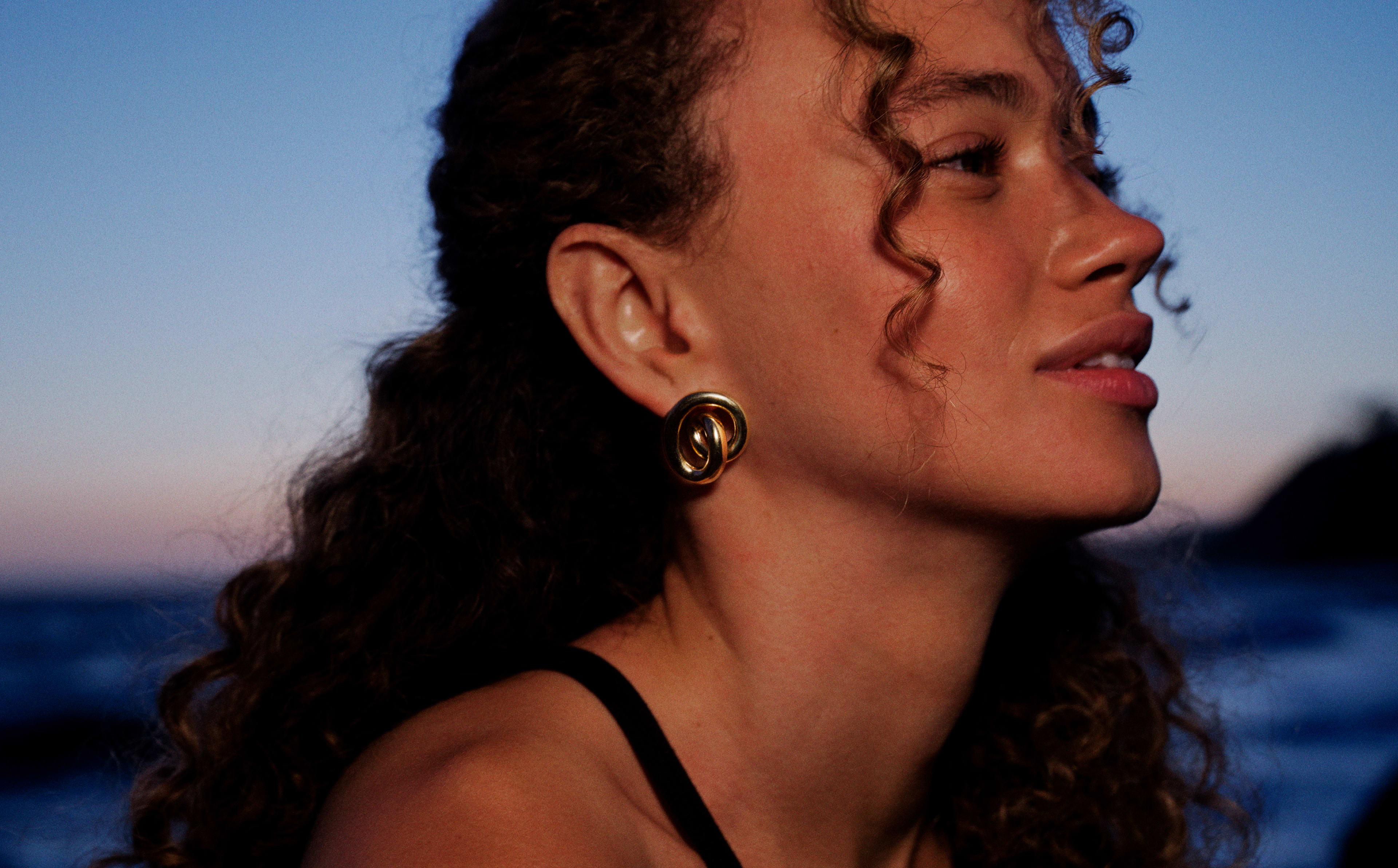 Woman in profile wearing gold chunky hoop earrings by the ocean at golden hour