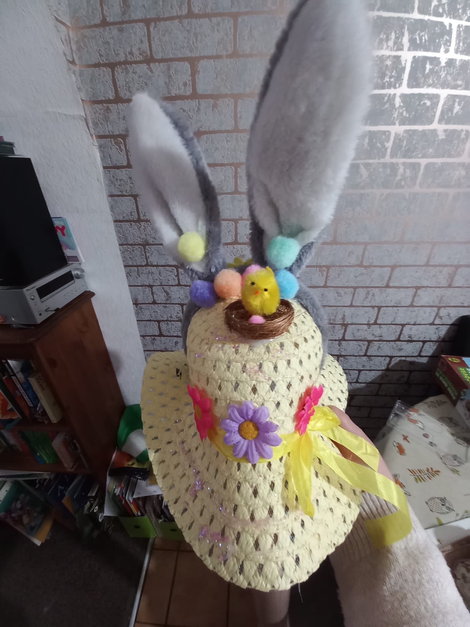 child' easter bonnet, straw coloured polyester woven fibres with grey rabbit ears headband and floral decorations, with a yellow ribbon and on top pompoms in various colours and a fibre nest with chick in it