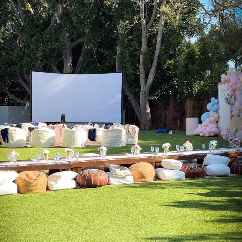 Garden celebration with a movie screen, white bean bags, a low dining table, and balloon decorations