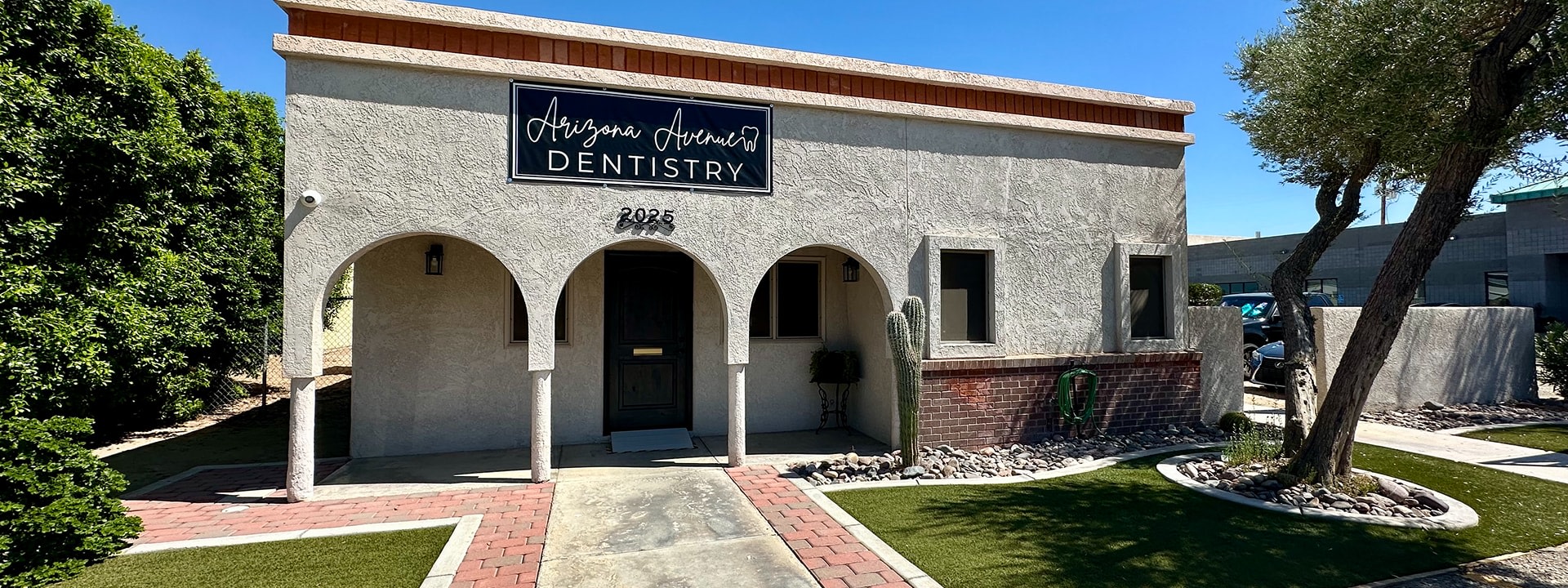 The Front entrance to the Arizona Avenue Dentistry office.  A while building with three arches and a sign with their logo above the street address 2025