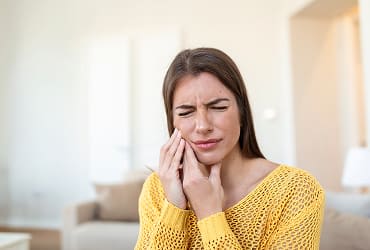 A woman in a yellow sweater holding her face as she is obviously in pain from a dental emergency she needs help with.