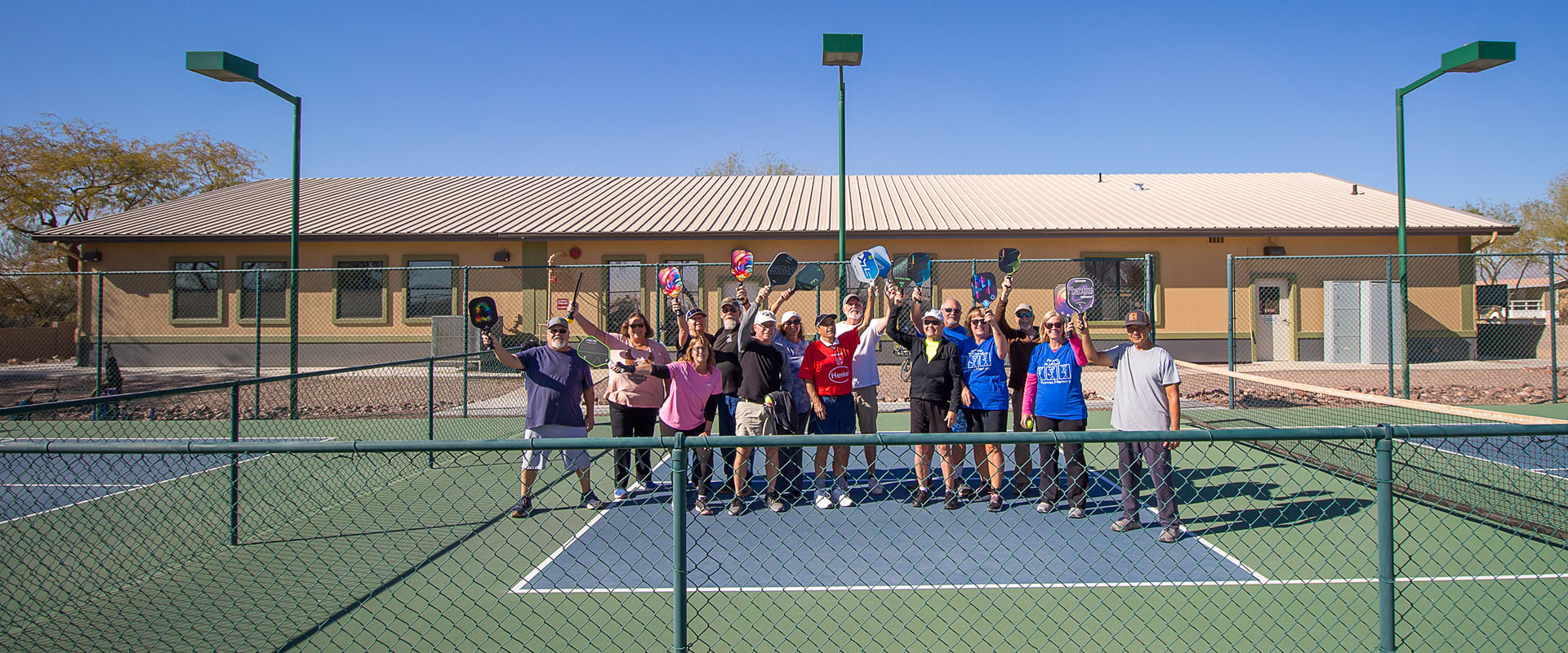 Group of active adults 55+ waving at the camera on a pickleball court at Cotton Lane RV Resort in Goodyear, AZ.