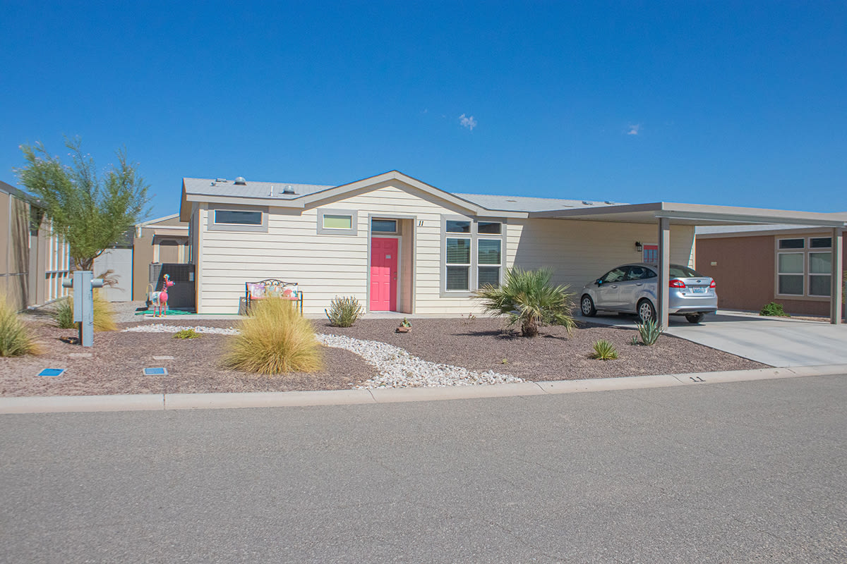 Light manufactured home with a bright pink front door, ornamental grasses and a car under the carport