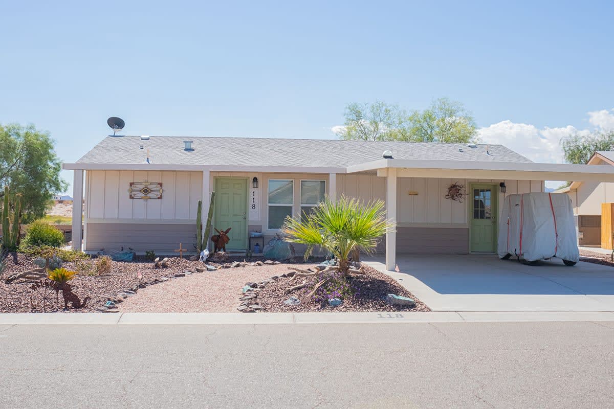 Pale manufactured home with sage green doors, a young palm and a cactus garden out front