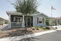 Blue grey manufactured home with a white porch railing, red shutters, shade tree and flagpole