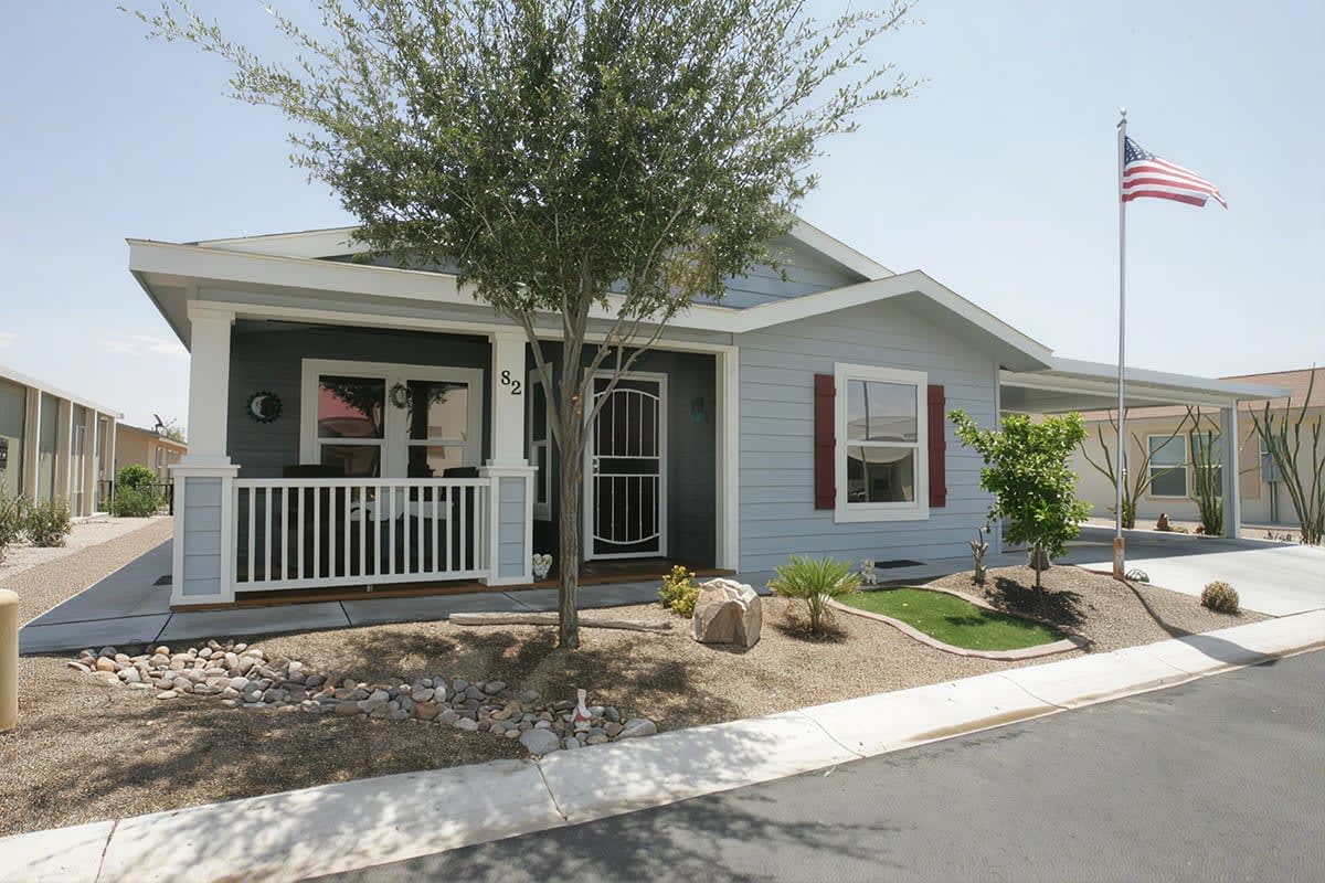 Blue grey manufactured home with a white porch railing, red shutters, shade tree and flagpole