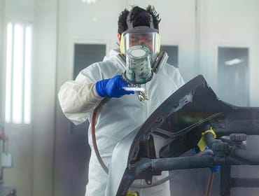 An employee in safety gear using a sprayer to apply paint to a body panel.