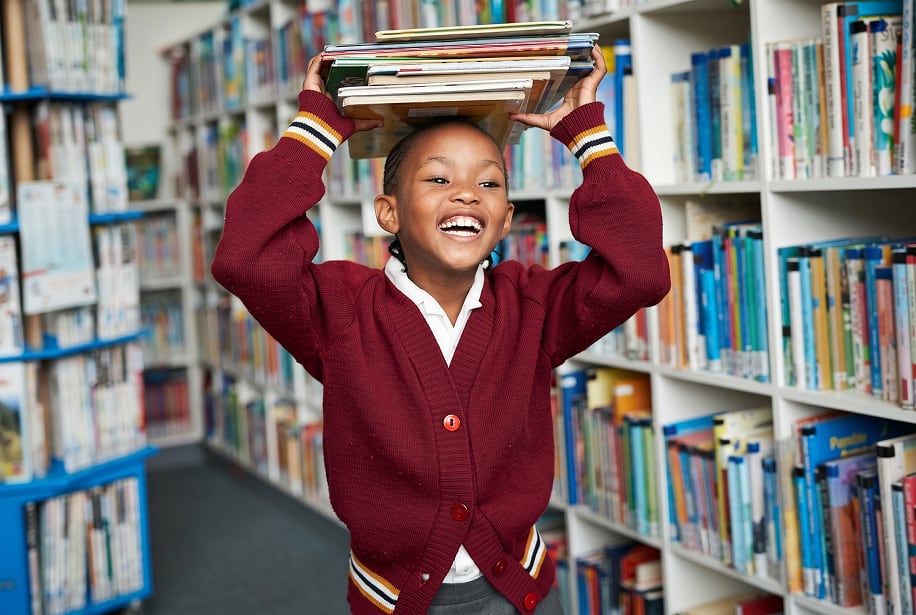 Smiling boy pupil balancing a stack of books on his head in a school library.