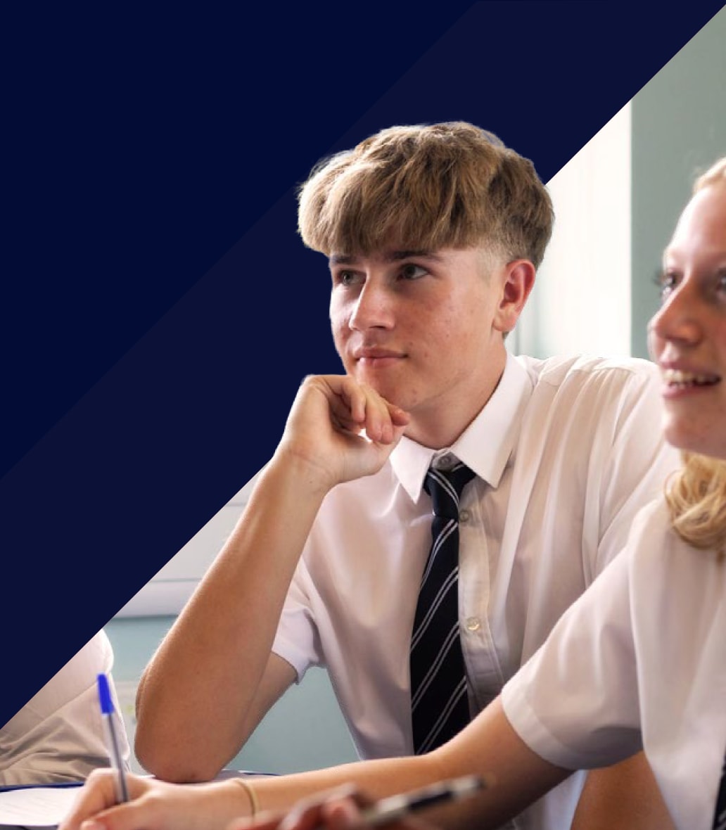 A secondary pupil in a white shirt and striped tie listens in class, chin on hand, beside a smiling classmate