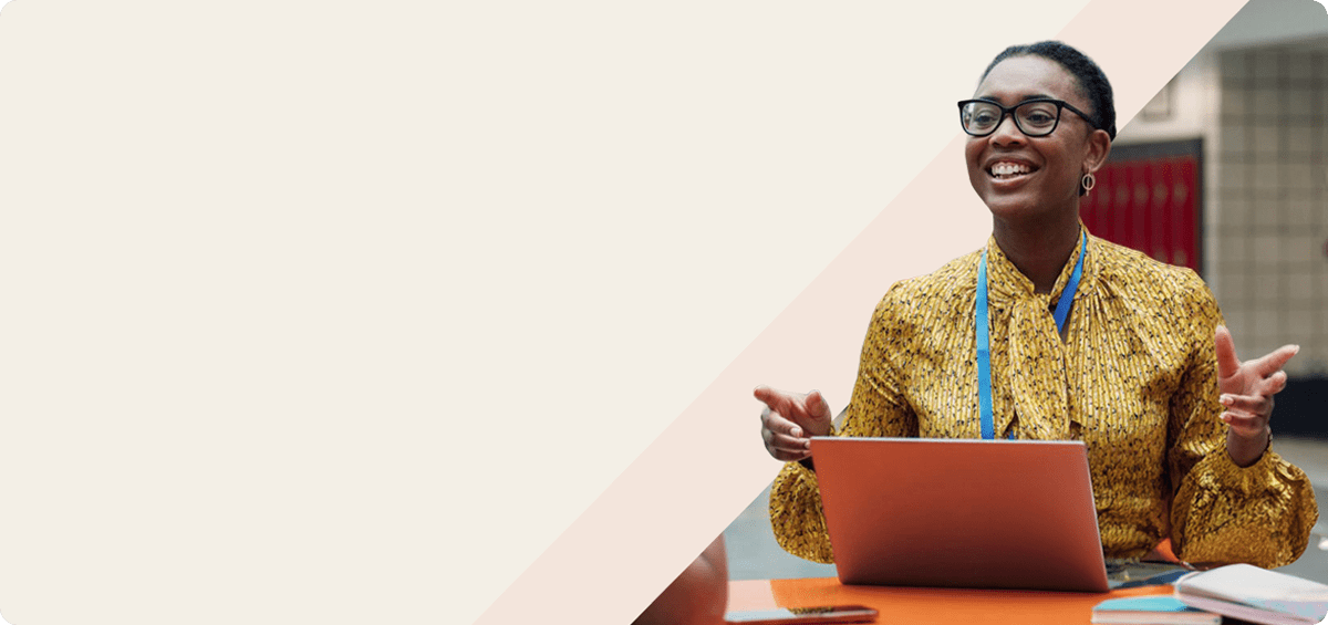 Smiling woman in glasses and a lanyard talks with open hands at a table with a laptop in a school building