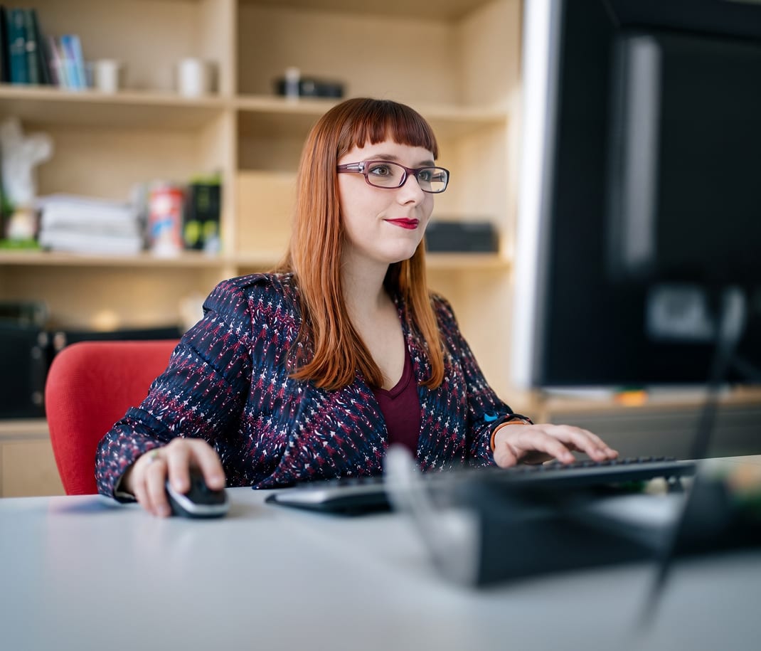 Woman with red hair and glasses working at a desktop computer in an office.