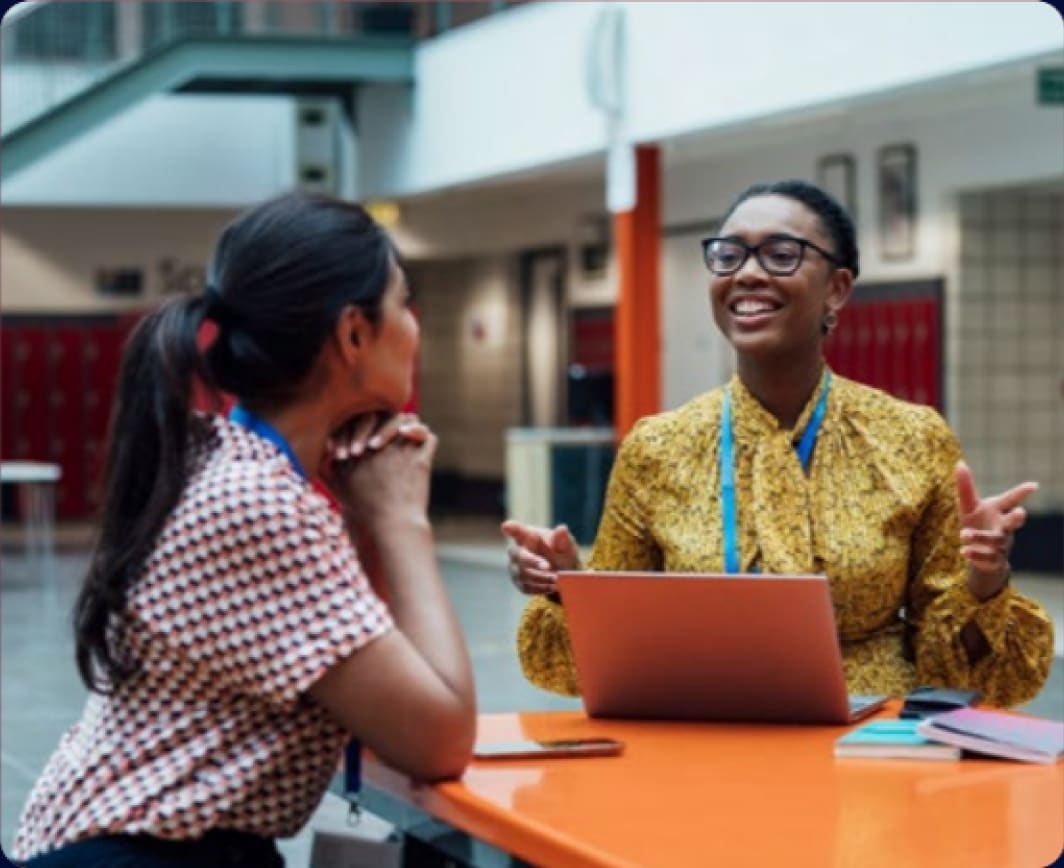 Two women talking across an orange table in a school atrium, one smiling beside a laptop
