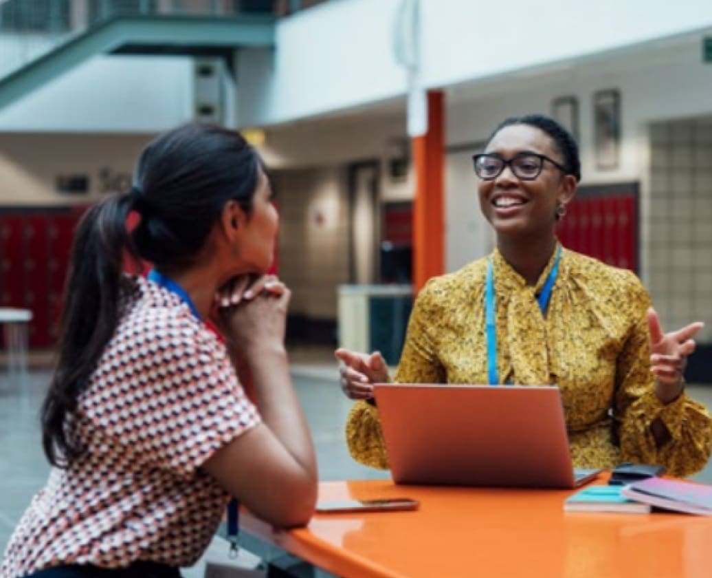 Two school staff members with lanyards talk at an orange table, one smiling and gesturing beside an open laptop