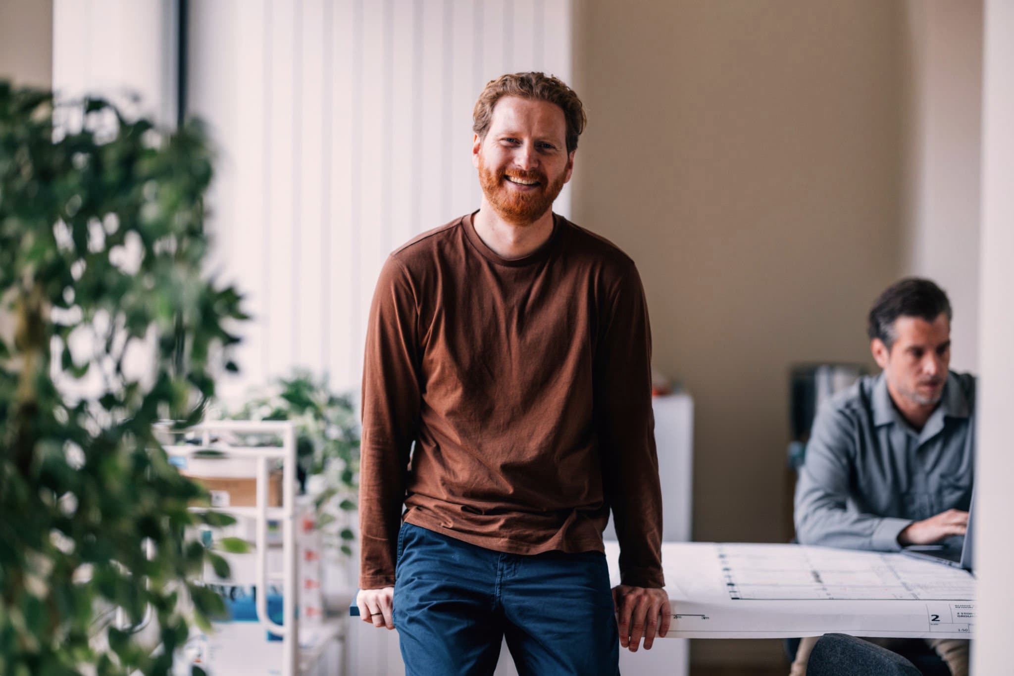 A smiling man leans on a table in a bright office while another man works on a laptop behind him