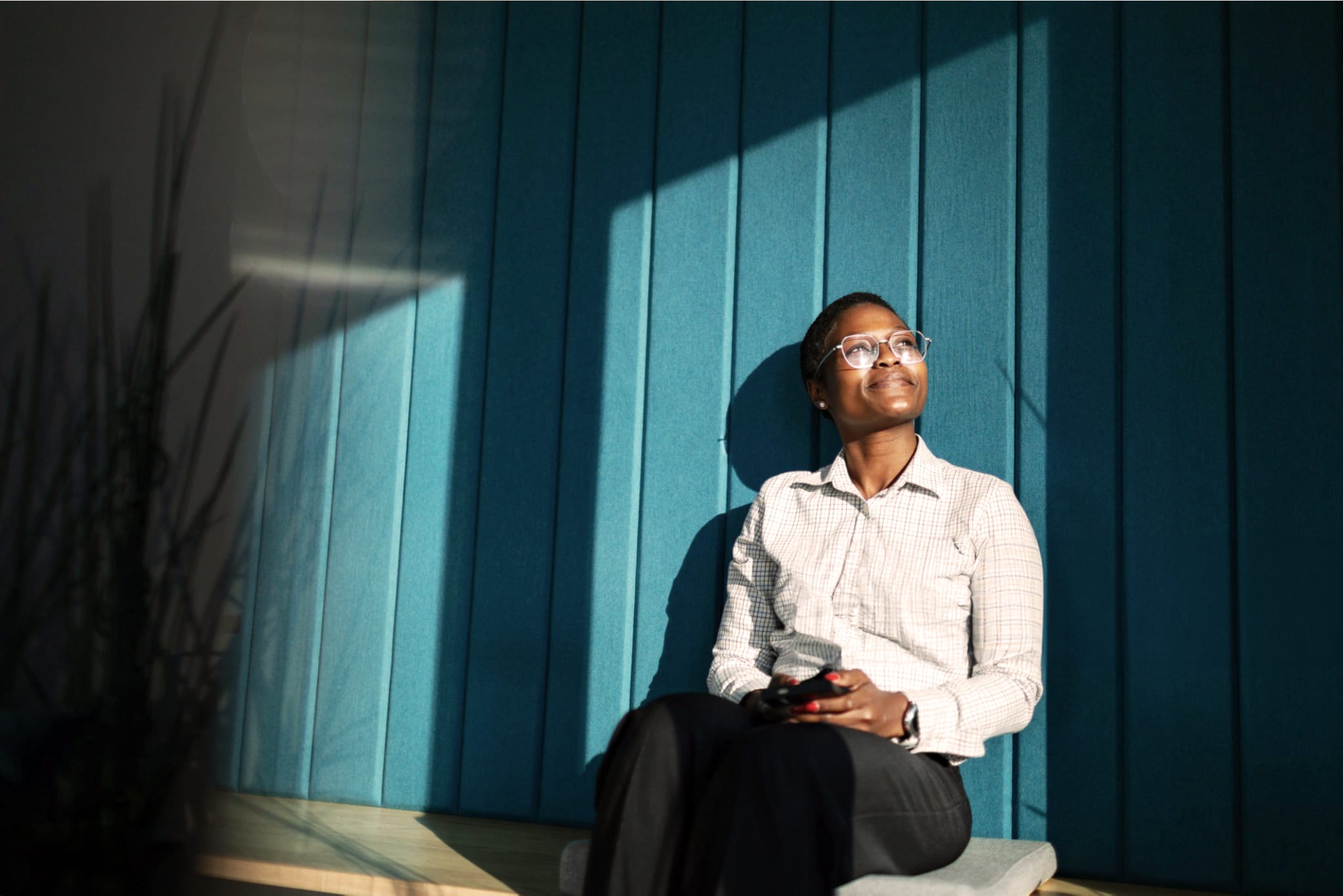 A woman wearing glasses sits in sunlight against a teal wall, looking upwards with a thoughtful smile.