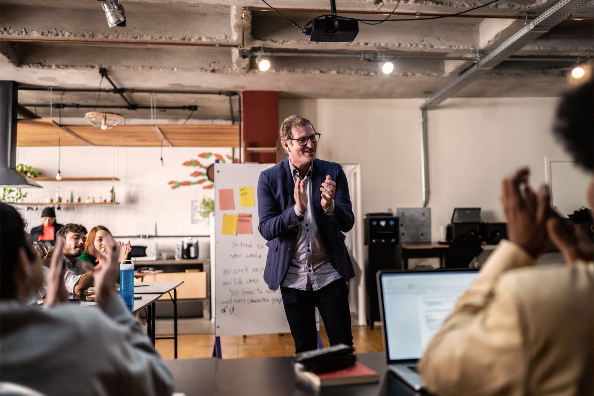 A man in a navy blazer presents to applauding colleagues in a modern office.