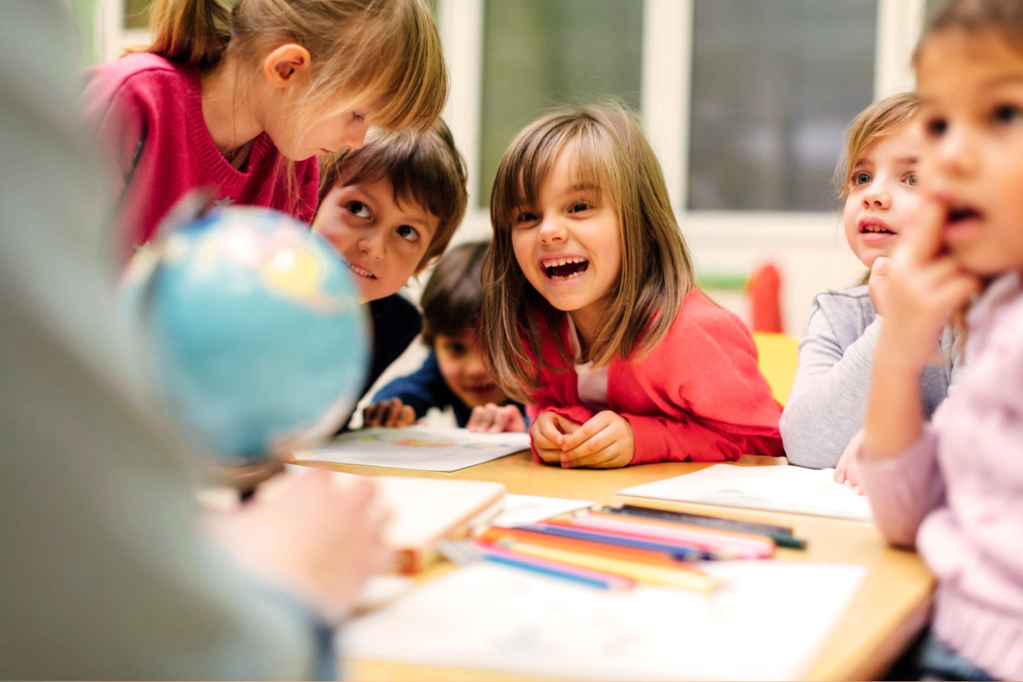 Young pupils smiling and chatting around a classroom table with a globe, worksheets and colouring pencils
