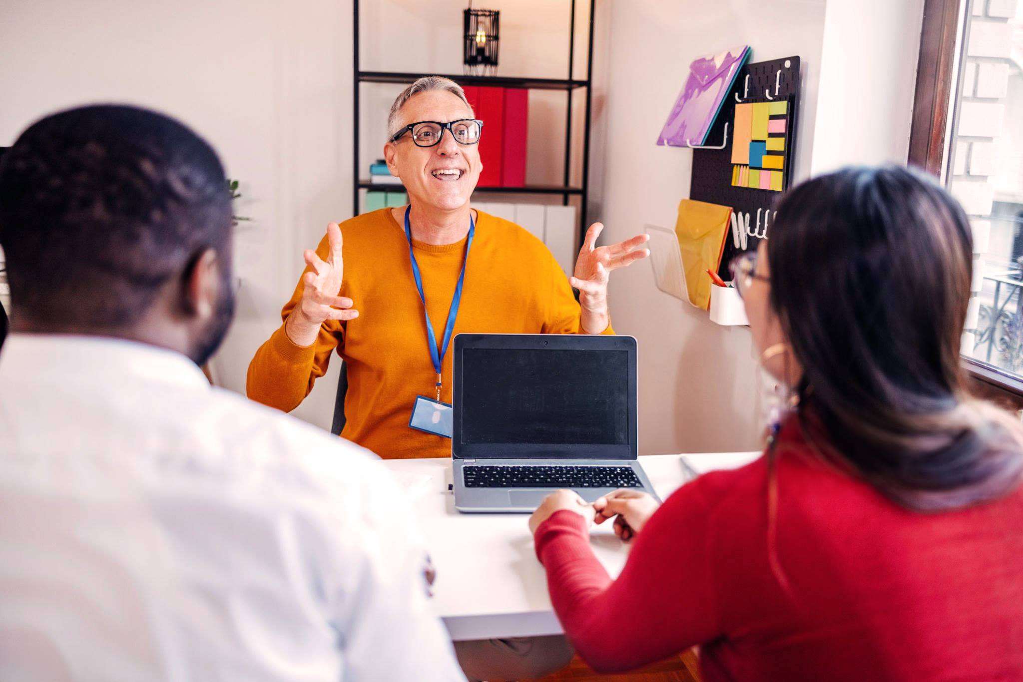 A smiling staff member with a lanyard and laptop talks with two colleagues across a desk