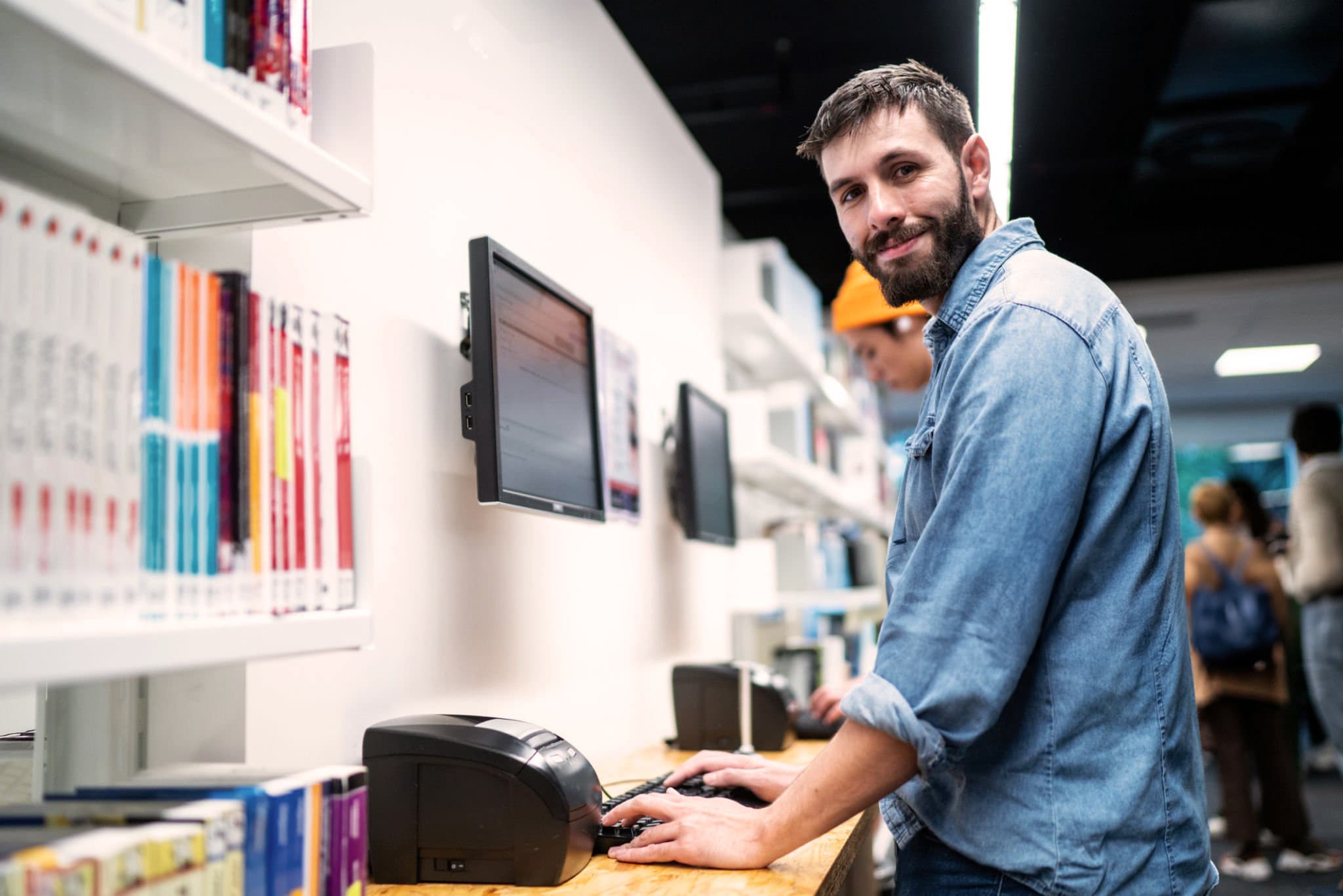 Smiling man in a denim shirt typing at a wall-mounted computer station beside library bookshelves